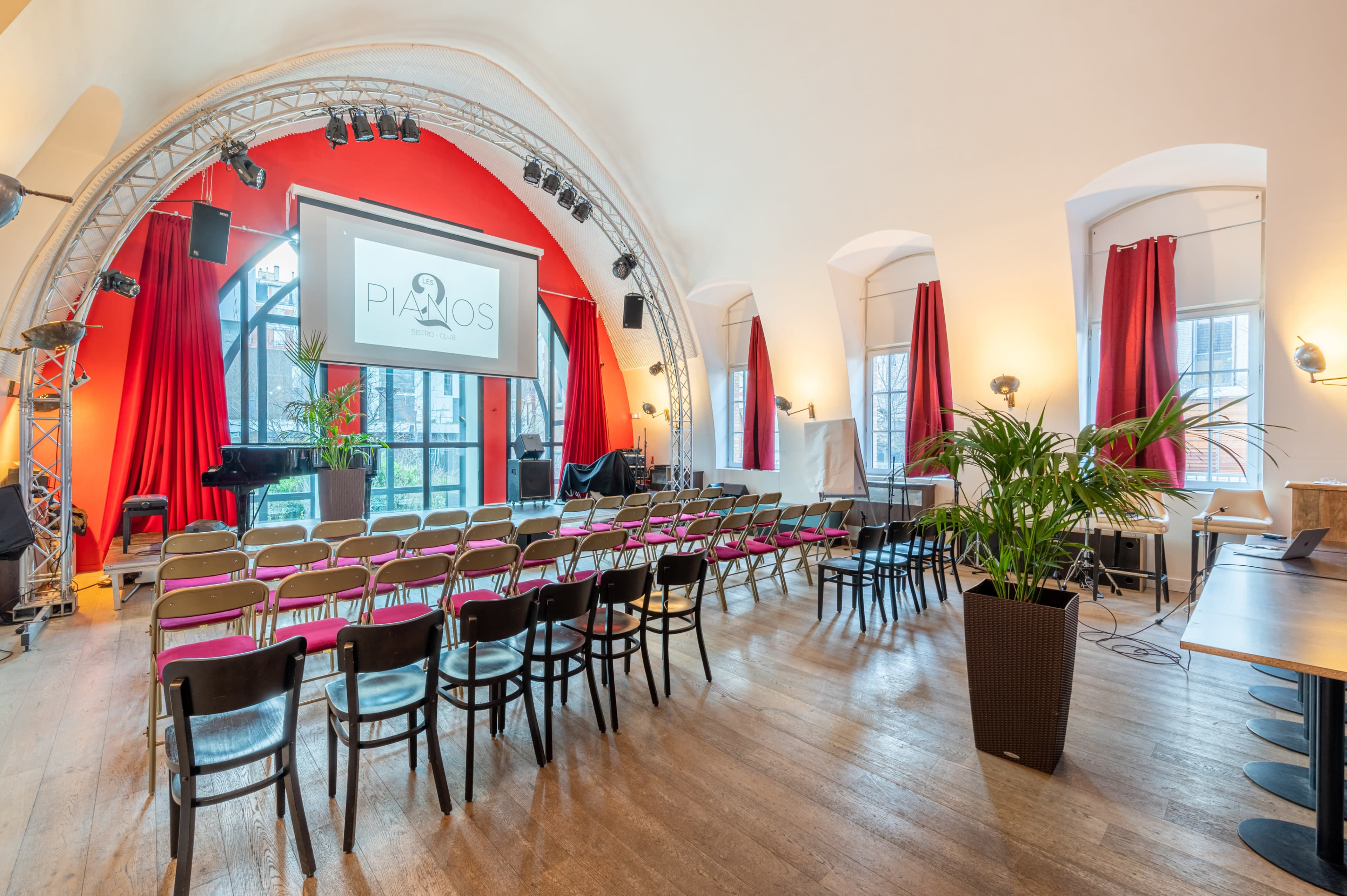 An indoor venue set up for an event, with rows of chairs facing a screen framed by red curtains and decorated with a piano.