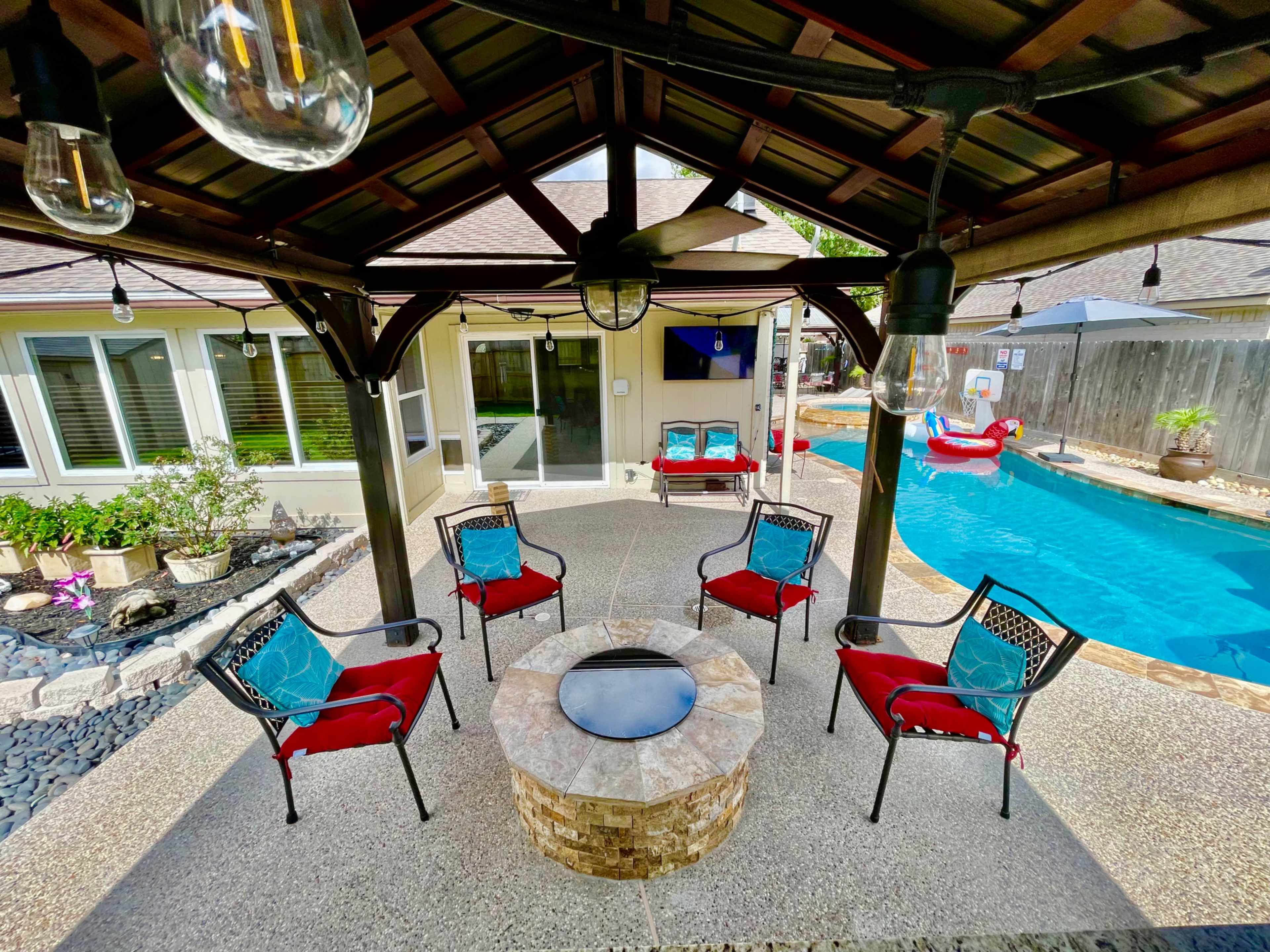 A covered patio features six red chairs arranged around a stone fire pit, with a swimming pool visible in the background.