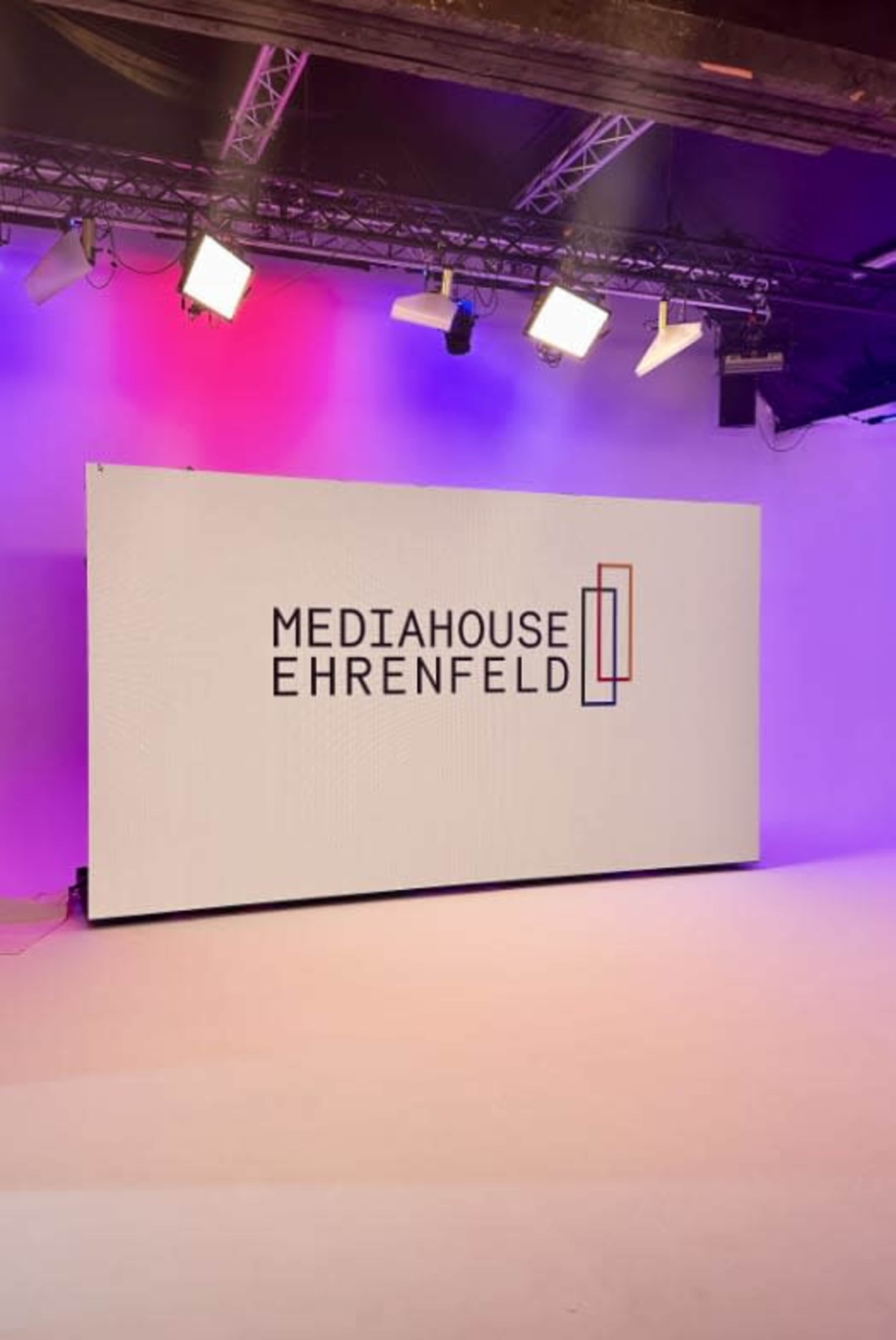 The image shows a large screen displaying the logo for "MEDIAHOUSE EHRENFELD" in a brightly lit studio setting.