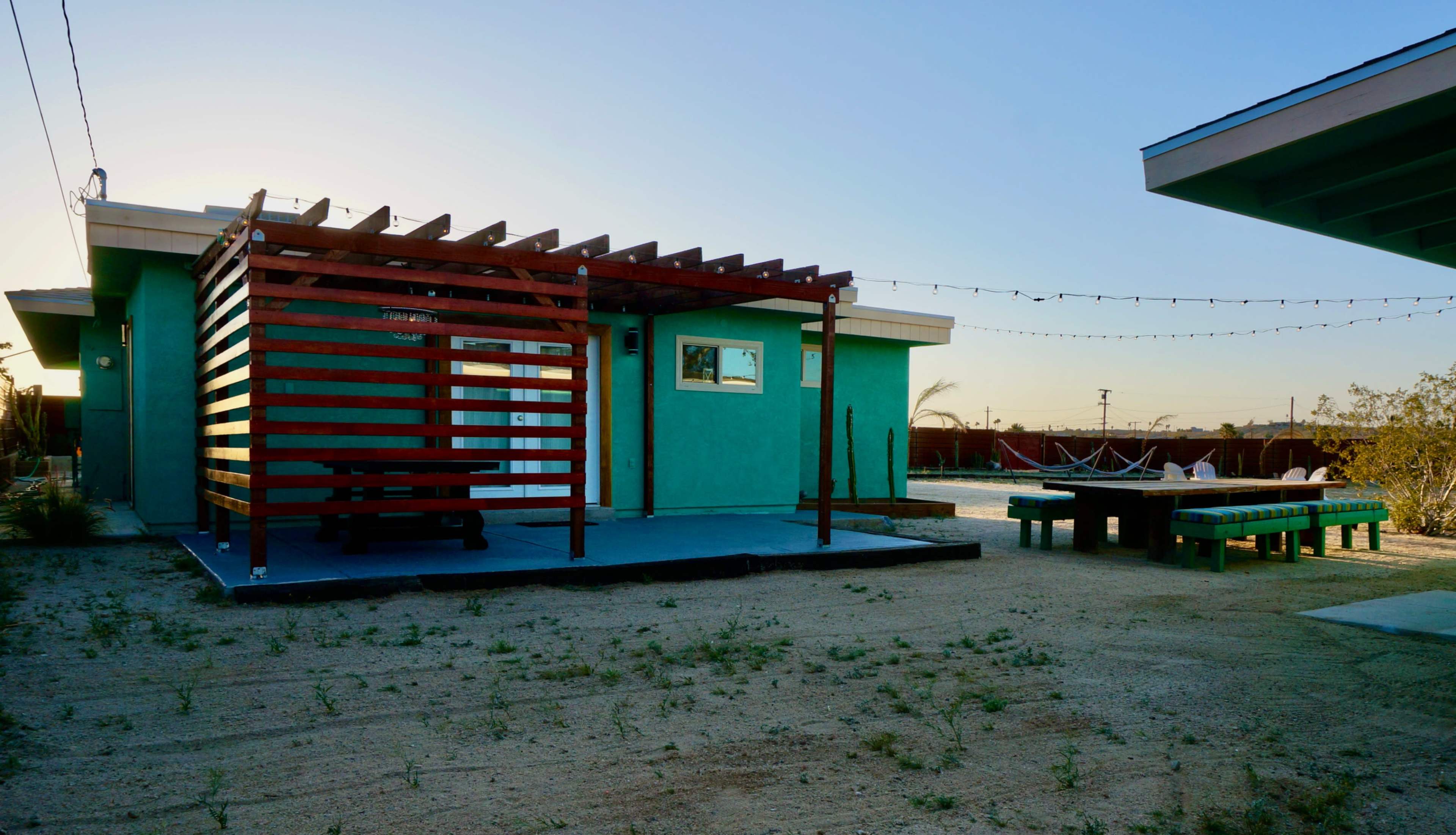 A small, brightly colored house with a wooden pergola and a gravel yard features a picnic table and a hammock in the background.