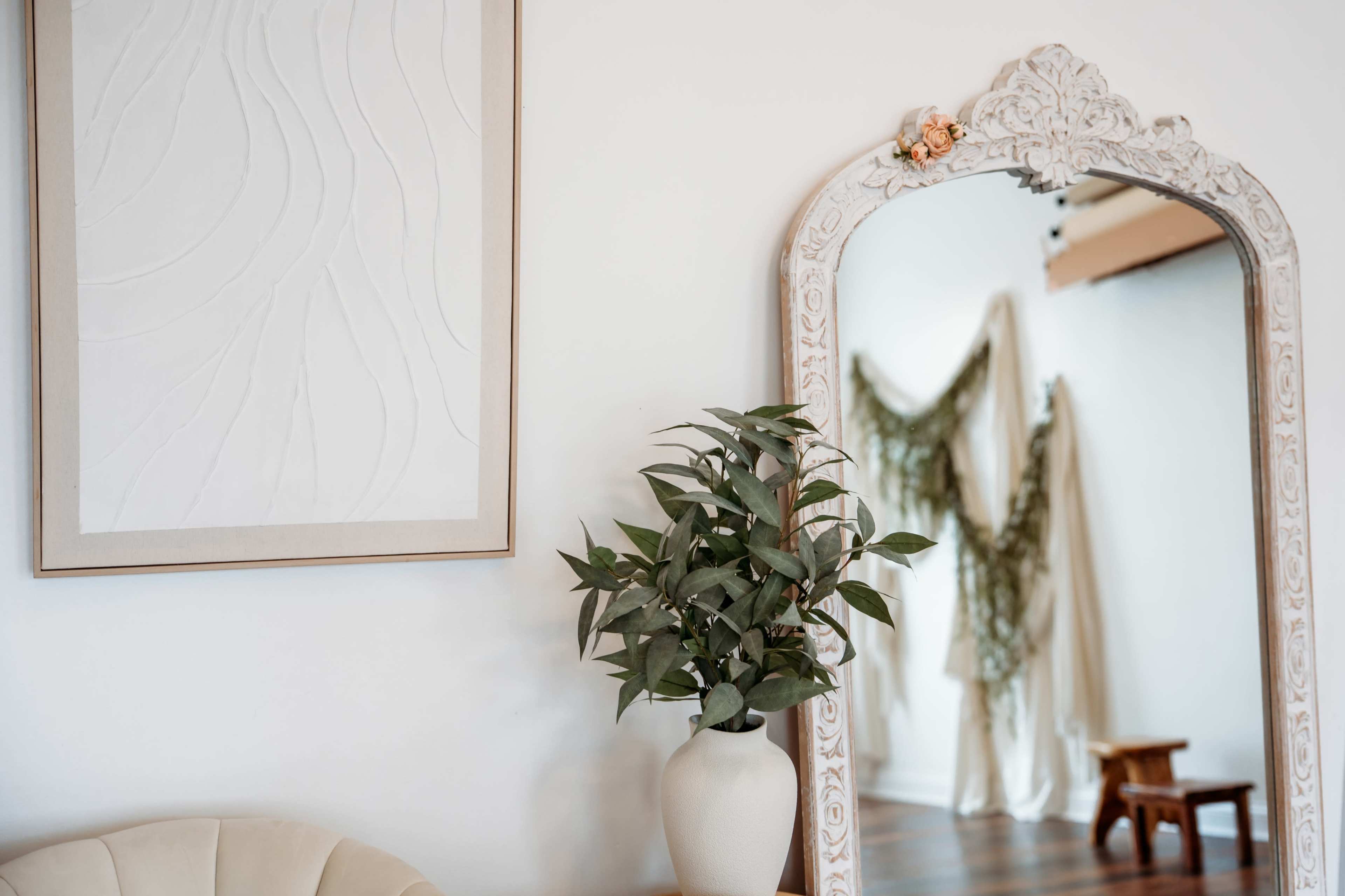 The image features a decorative mirror next to a potted plant, with a minimalist wall art piece above and soft fabric drapes in the background.