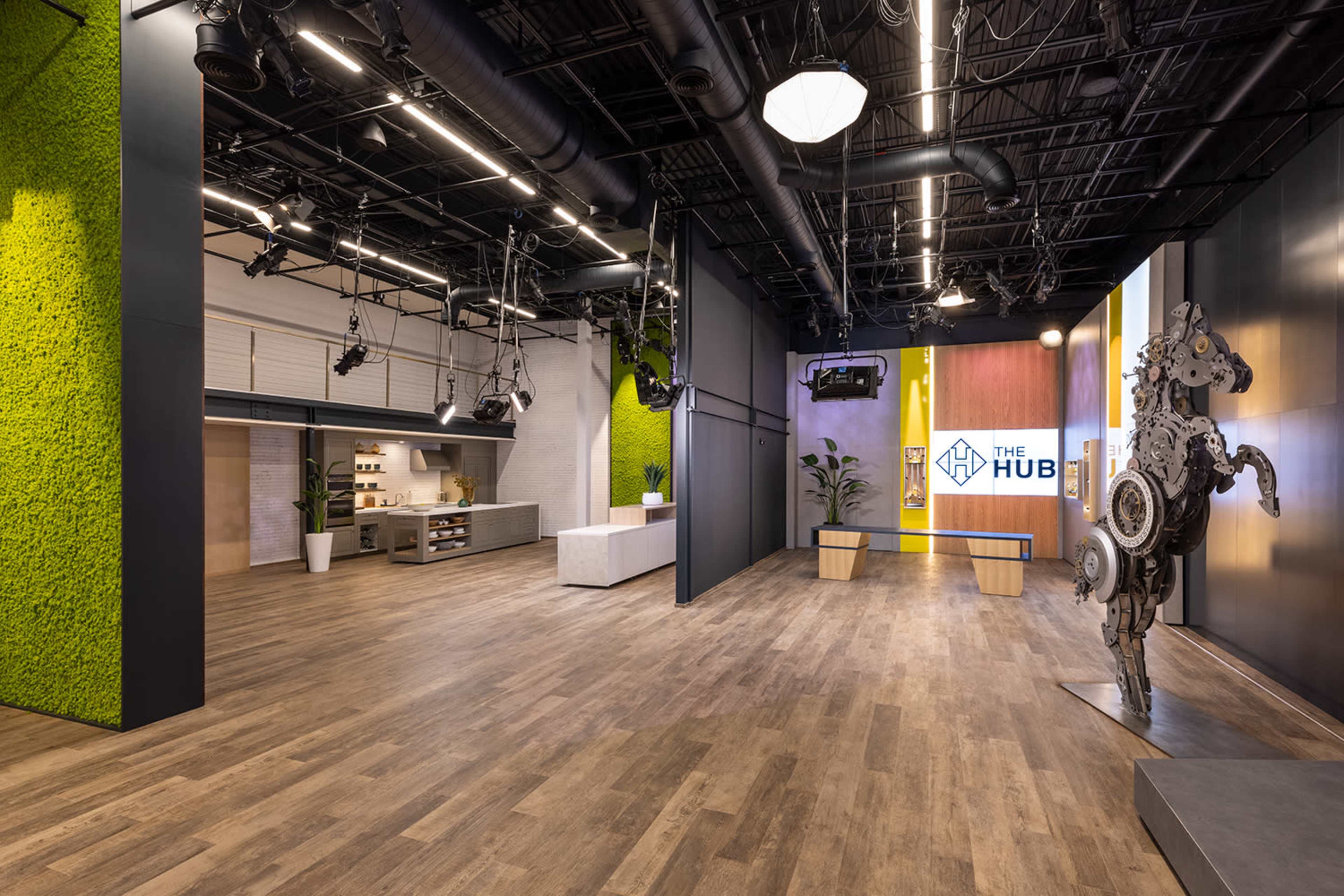 The image shows a modern, open interior space featuring wooden flooring, green moss-covered walls, and a display area with a large sign reading "THE HUB."