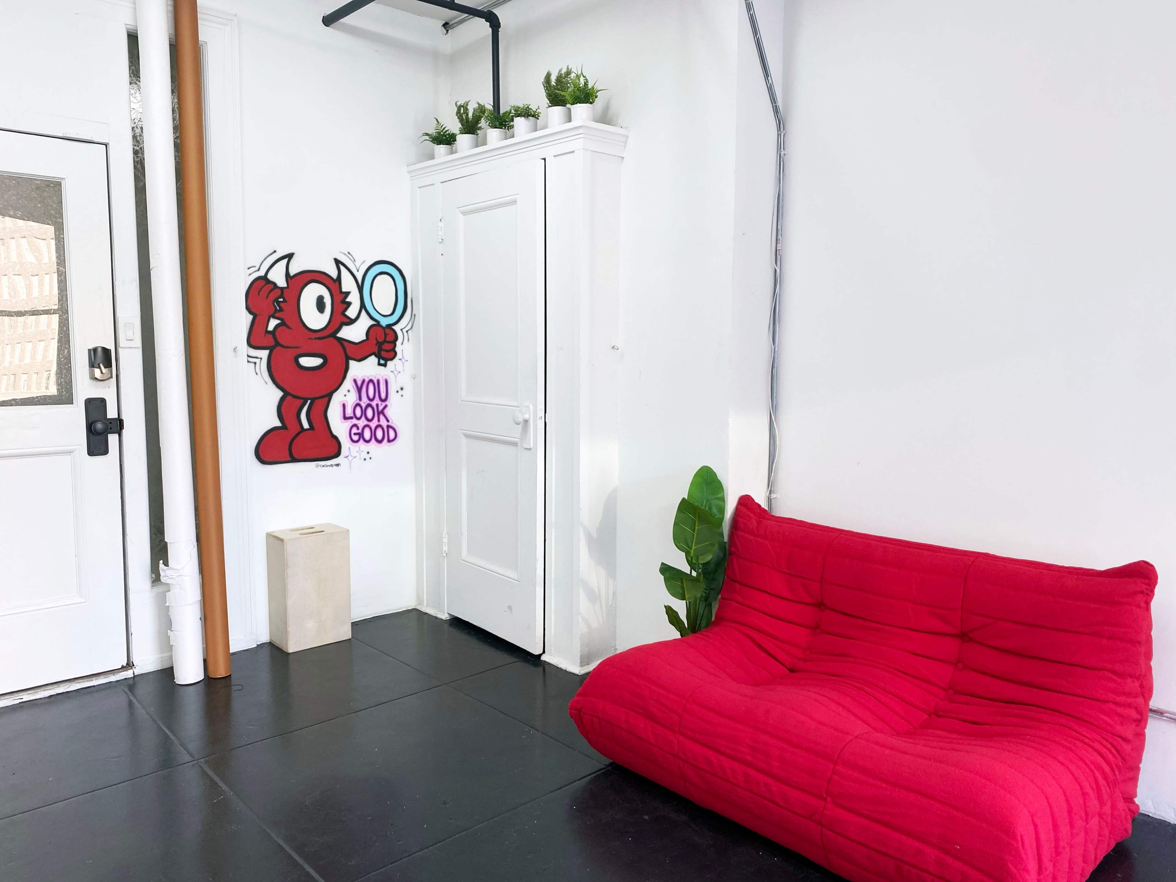 A bright, minimalist room with a red sofa, a white cabinet, a small plant, and a mural of a cartoon character with the phrase "YOU LOOK GOOD" on the wall.