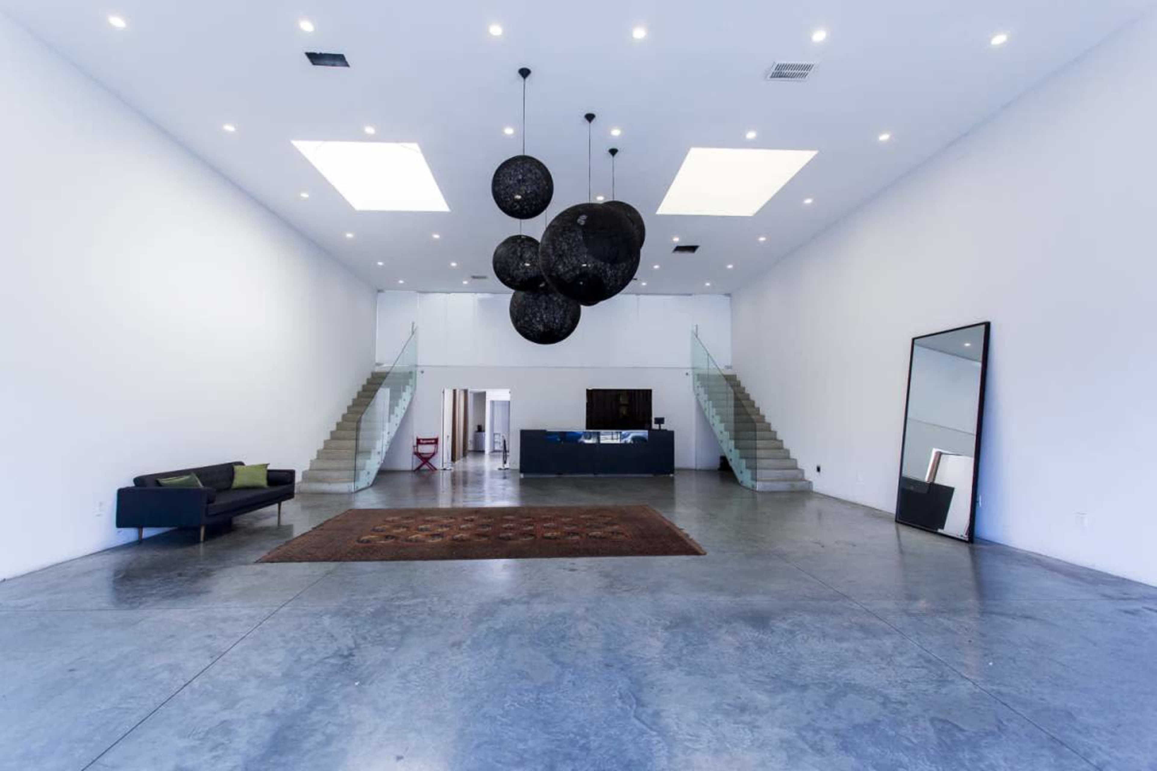 The image shows a spacious, minimalistic interior with polished concrete floors, a large area rug, and a series of hanging black light fixtures above a modern seating area and an open staircase.