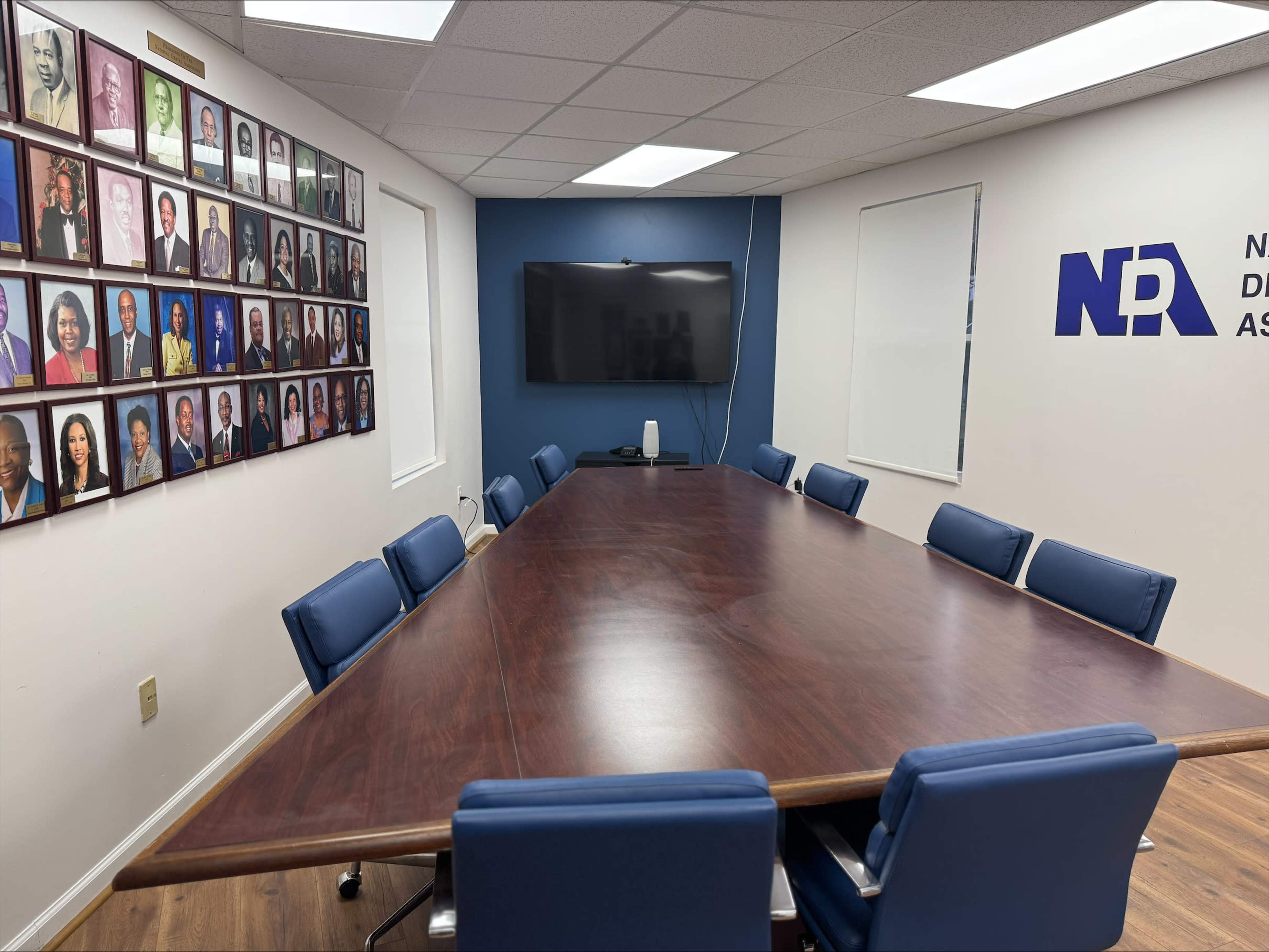 A conference room features a large wooden table surrounded by blue chairs, with a wall adorned by a collection of framed portraits and a television mounted on an opposite wall.