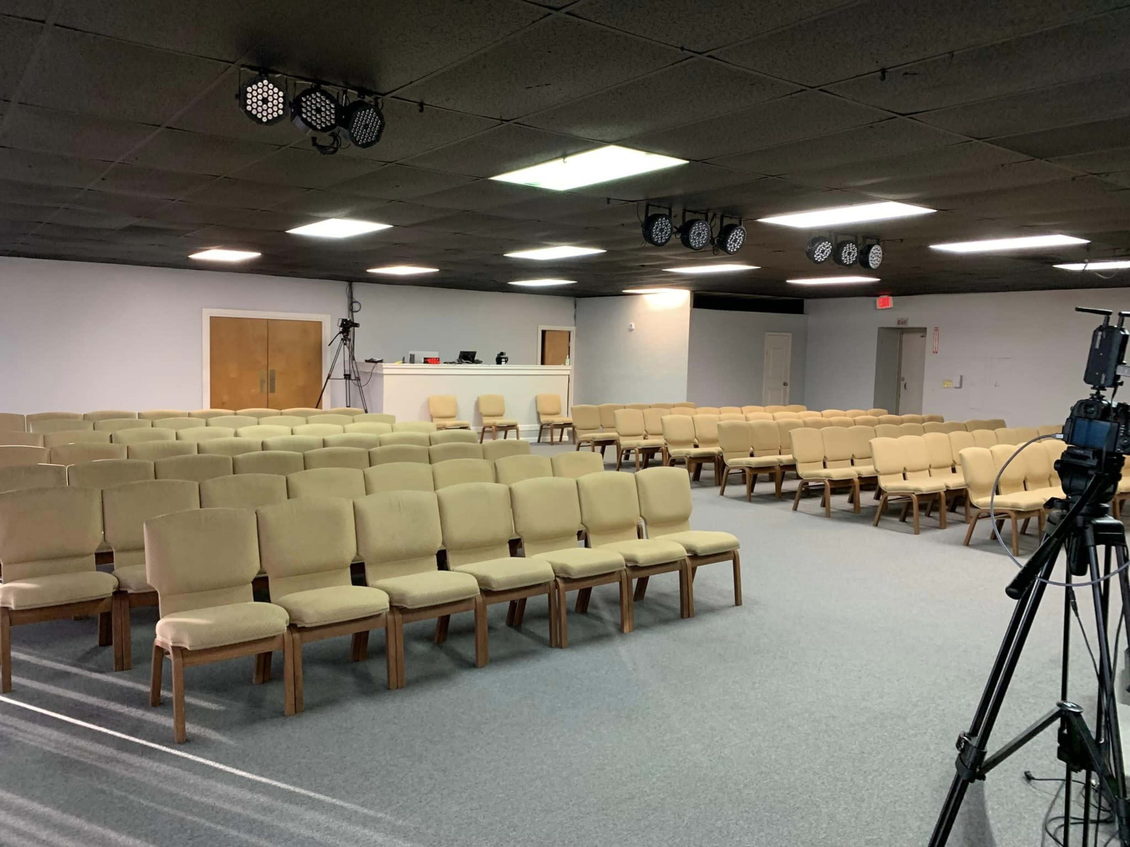 A spacious, empty room filled with rows of beige chairs set up for an event, with a camera and equipment positioned in the foreground.