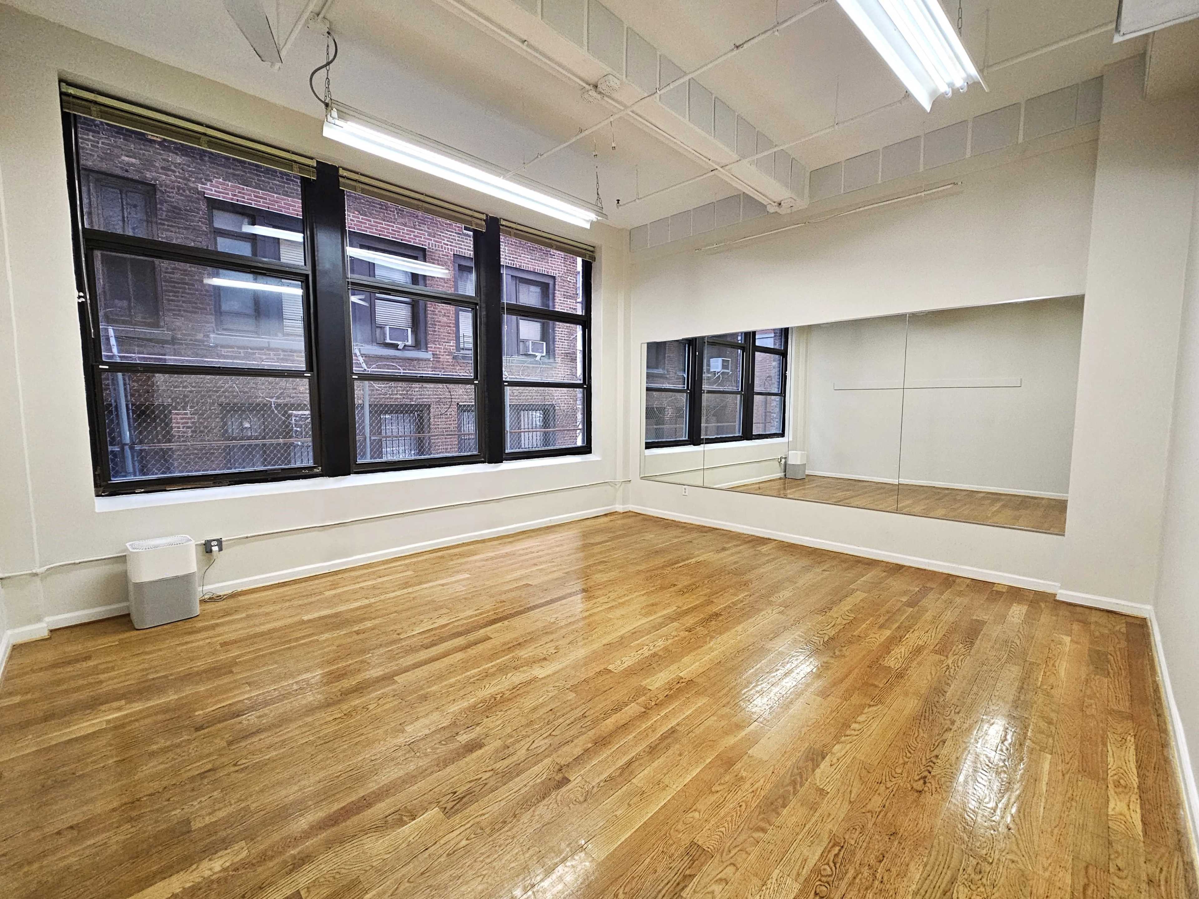 A spacious, empty room with wooden floors, large windows, and a wall-mounted mirror reflecting the interior.