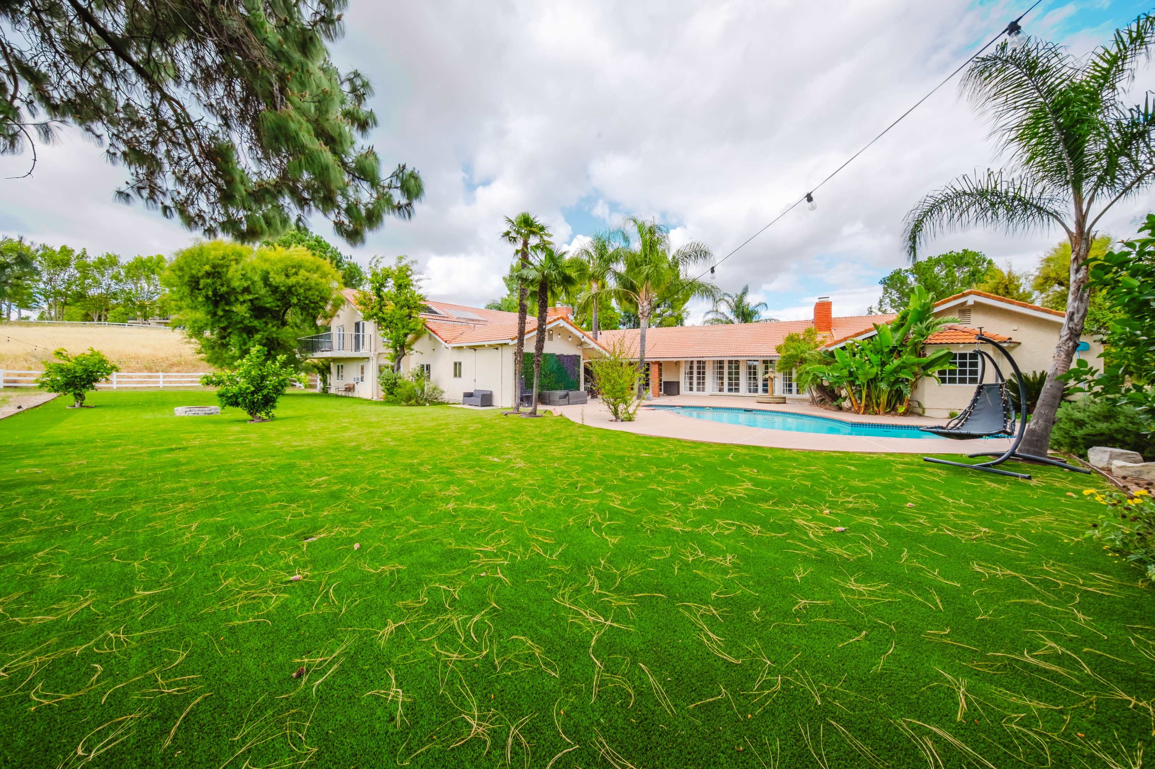 The image shows a spacious backyard featuring a well-maintained lawn, a swimming pool, and a house surrounded by palm trees and other greenery.