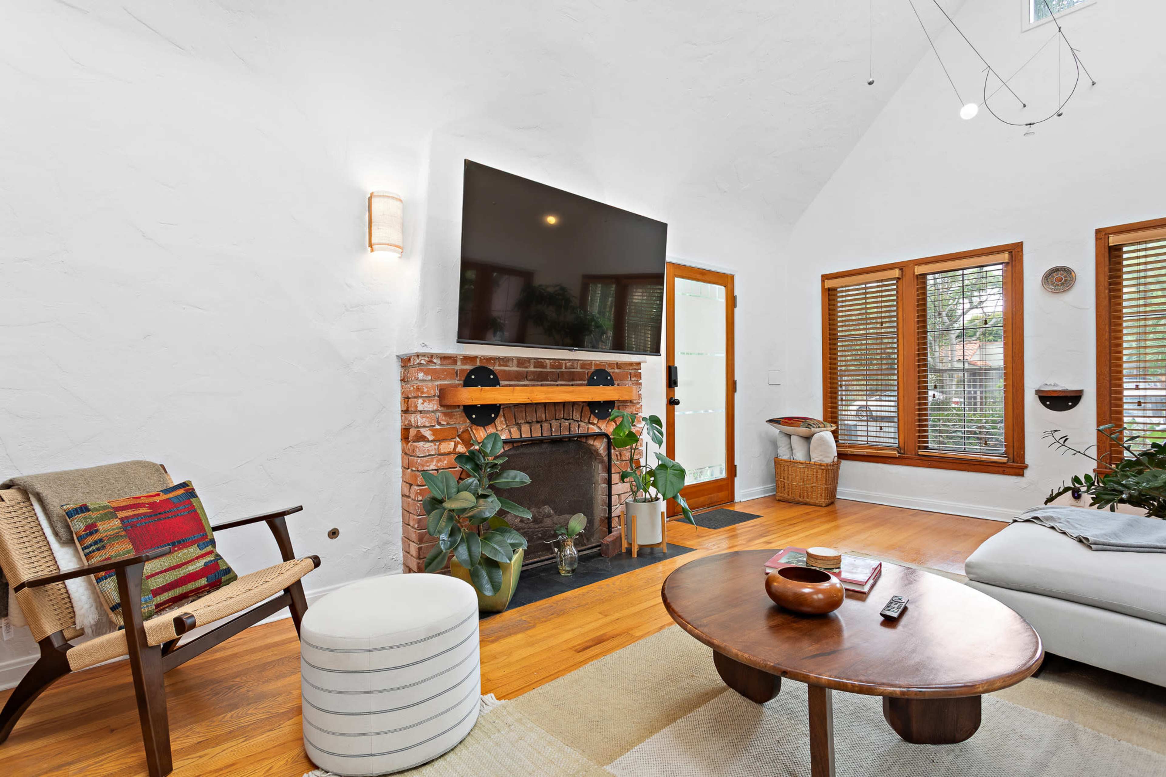 The image shows a bright living room featuring a brick fireplace, large television mounted on the wall, and modern furniture paired with indoor plants.