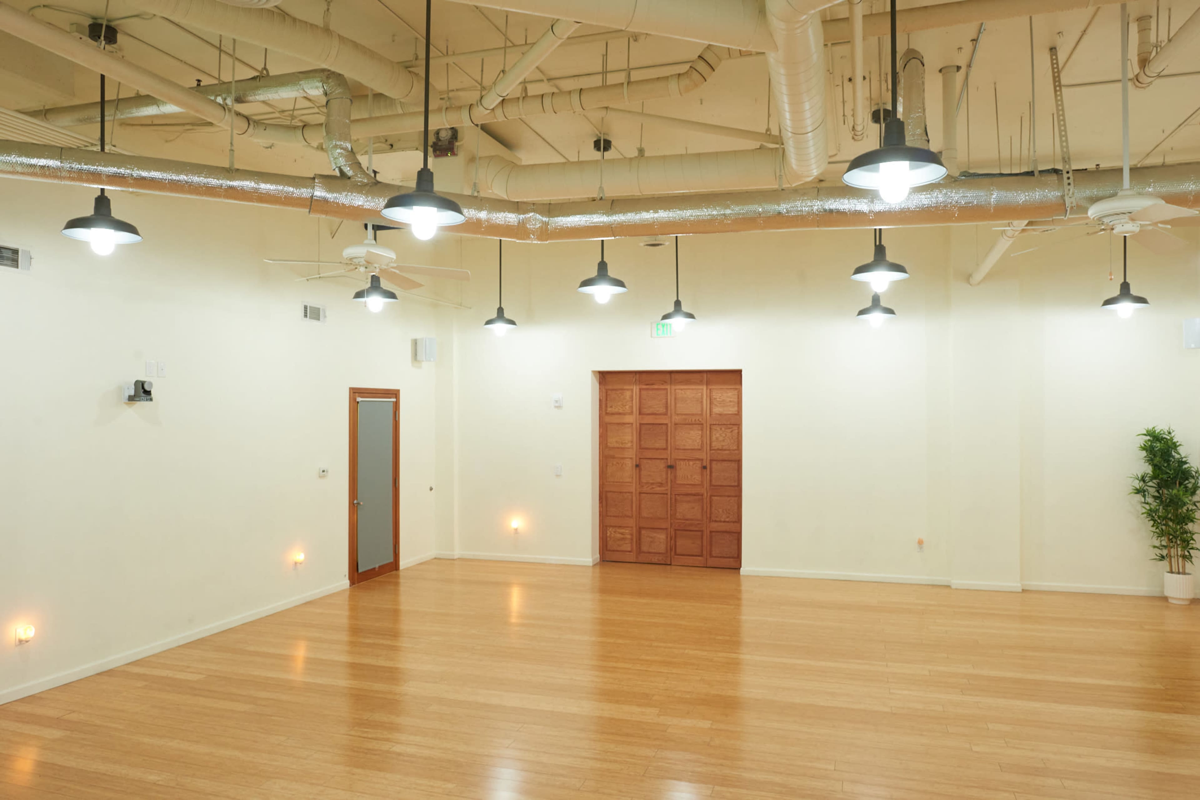 A spacious, well-lit interior room with wooden flooring, ceiling fans, and a large wooden door at one end.