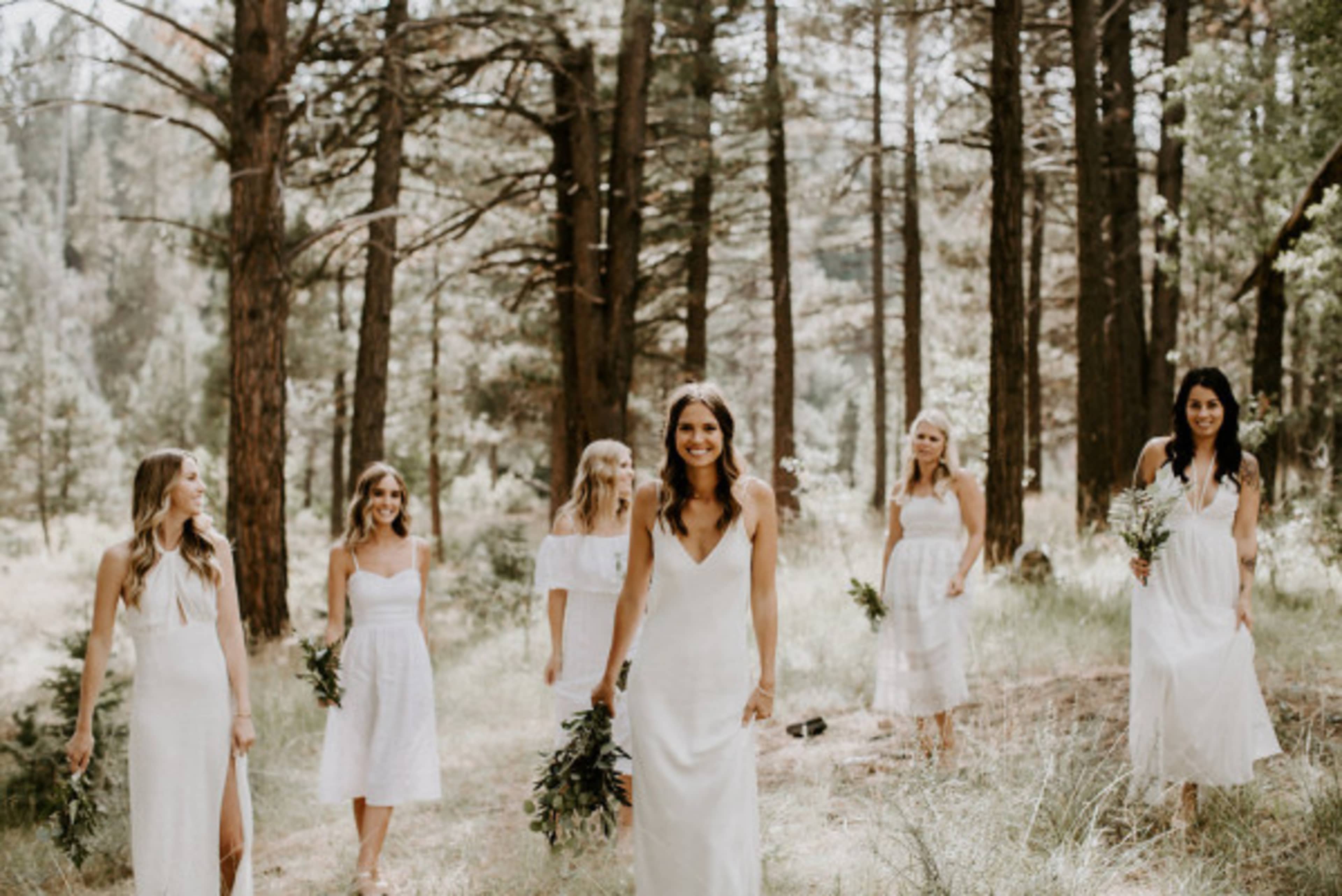 A group of six women wearing white dresses walks through a forested area.