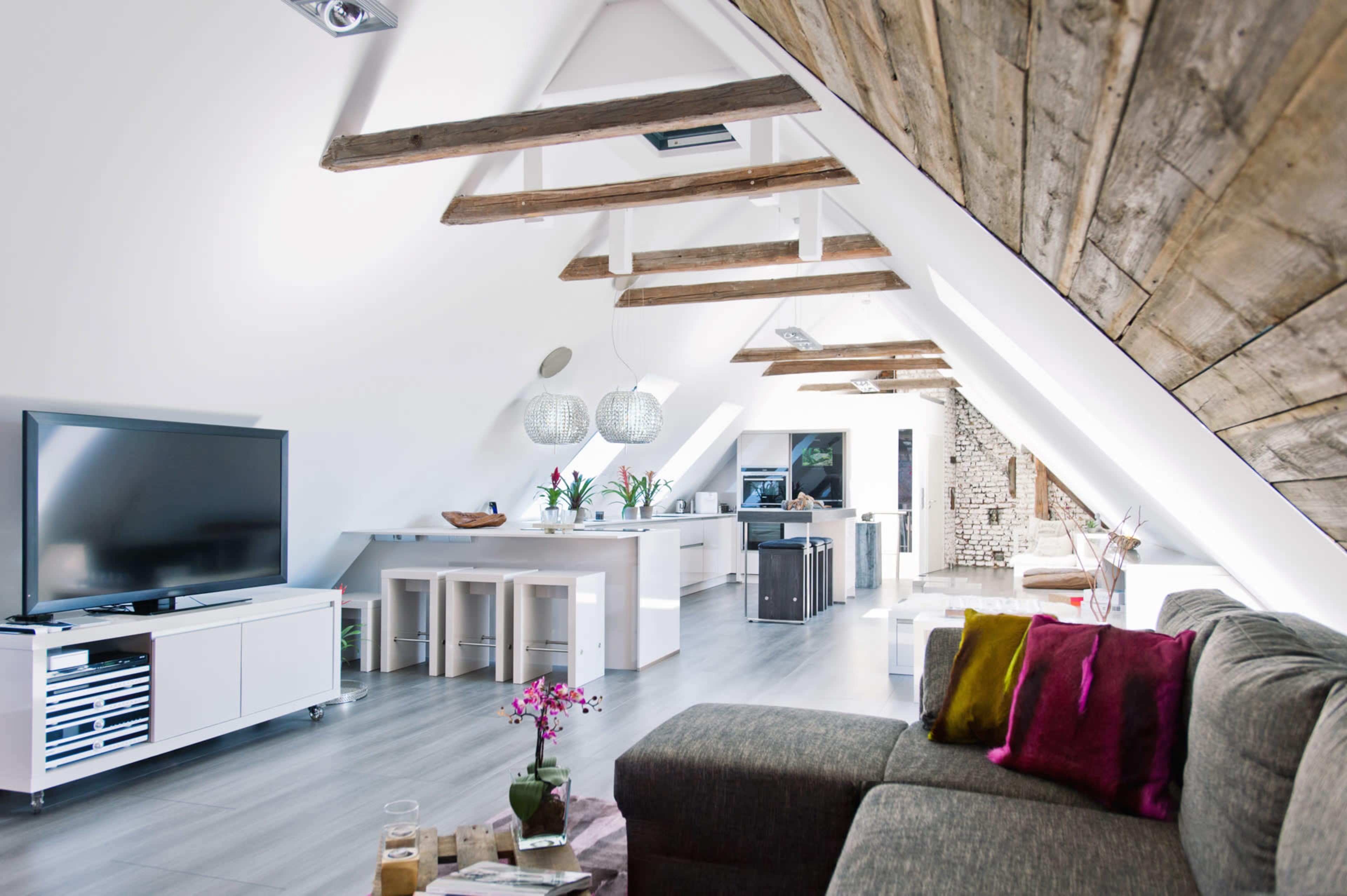 The image shows a modern open-plan attic living space featuring a large sofa, a television, a kitchen area with bar stools, and exposed wooden beams on the ceiling.