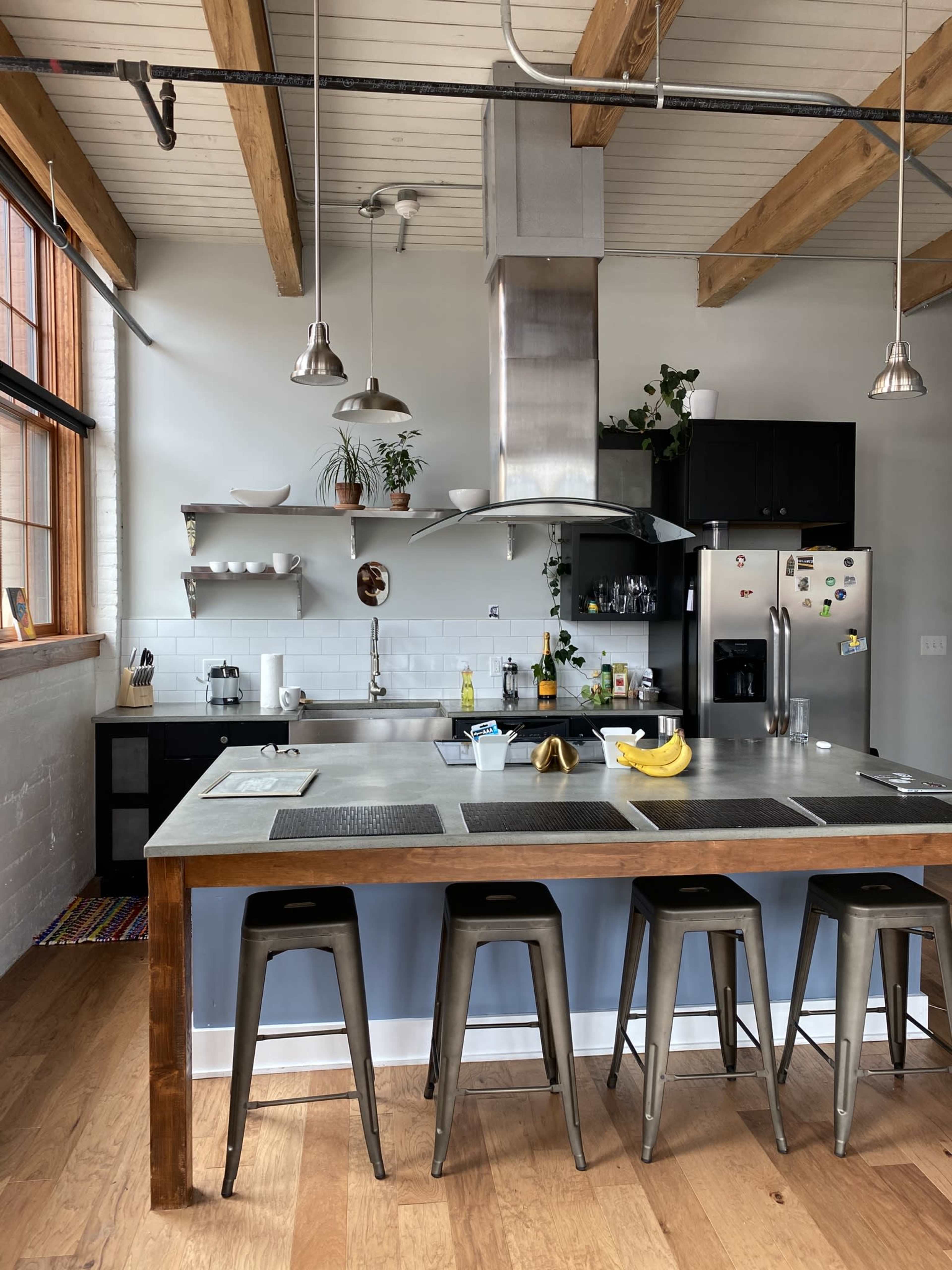 A modern kitchen with metal bar stools, a concrete counter, and stainless steel appliances, featuring a spacious layout and wooden beams overhead.