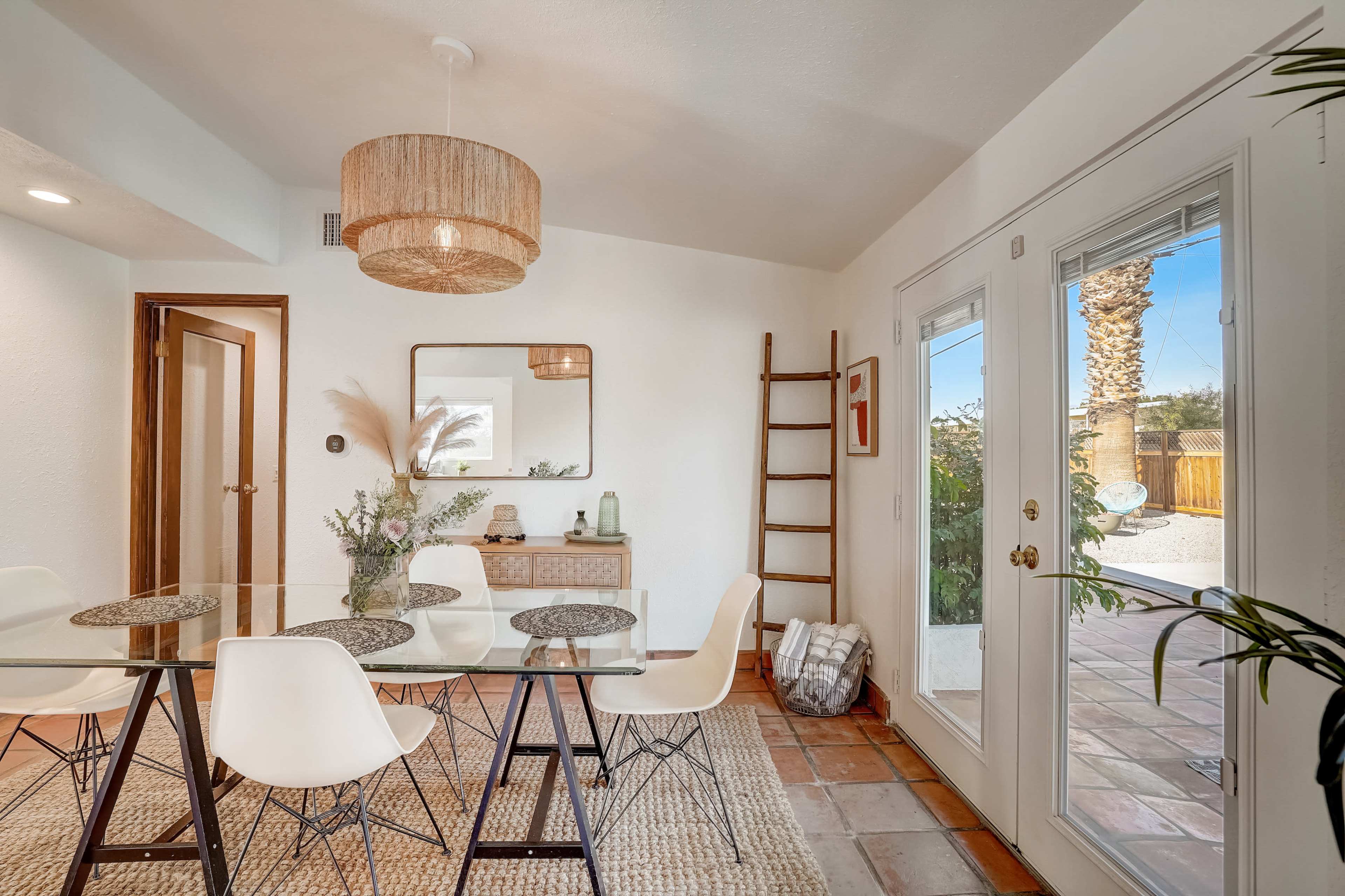 A dining area features a glass table surrounded by white chairs, with a woven light fixture above and doors opening to an outdoor space.