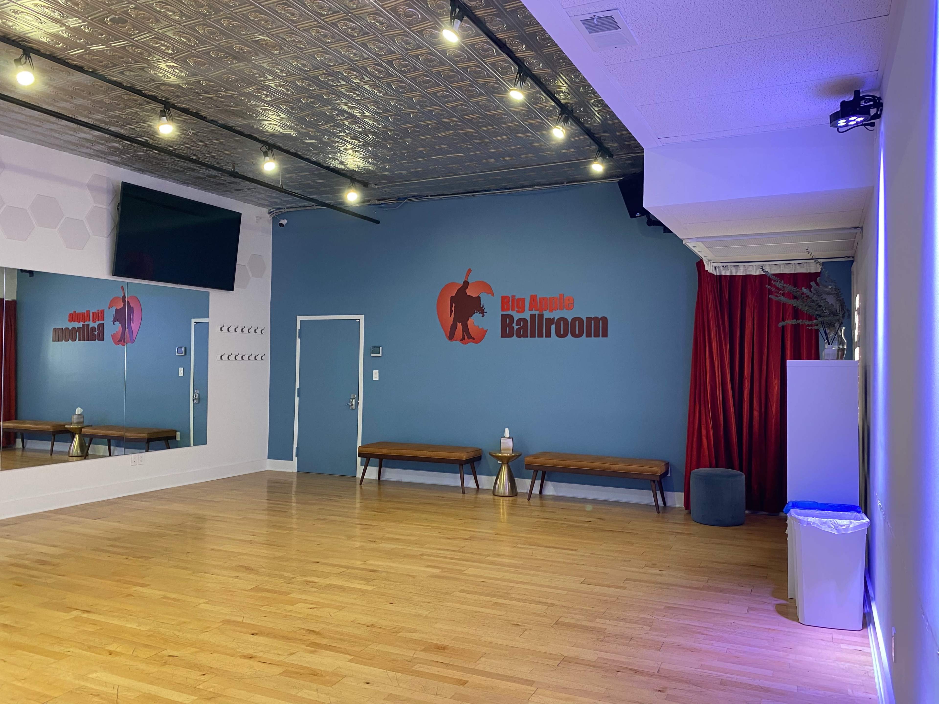 The image shows an empty dance studio with wooden flooring, a large mirror on one wall, a red curtain, and the words "Big Apple Ballroom" displayed on a blue wall.
