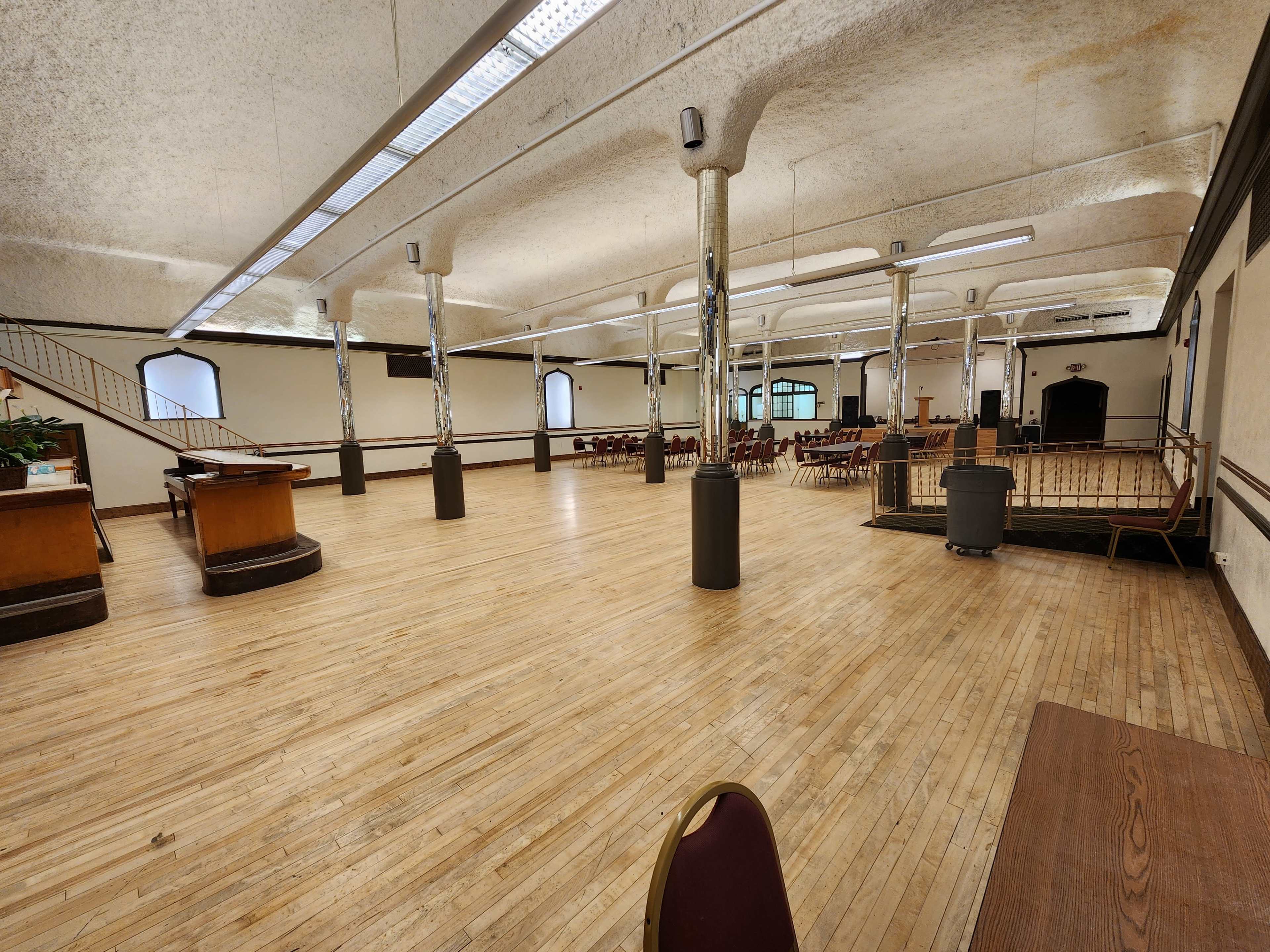 The image shows a spacious, empty hall with wooden flooring, pillars, and rows of chairs arranged in the background.