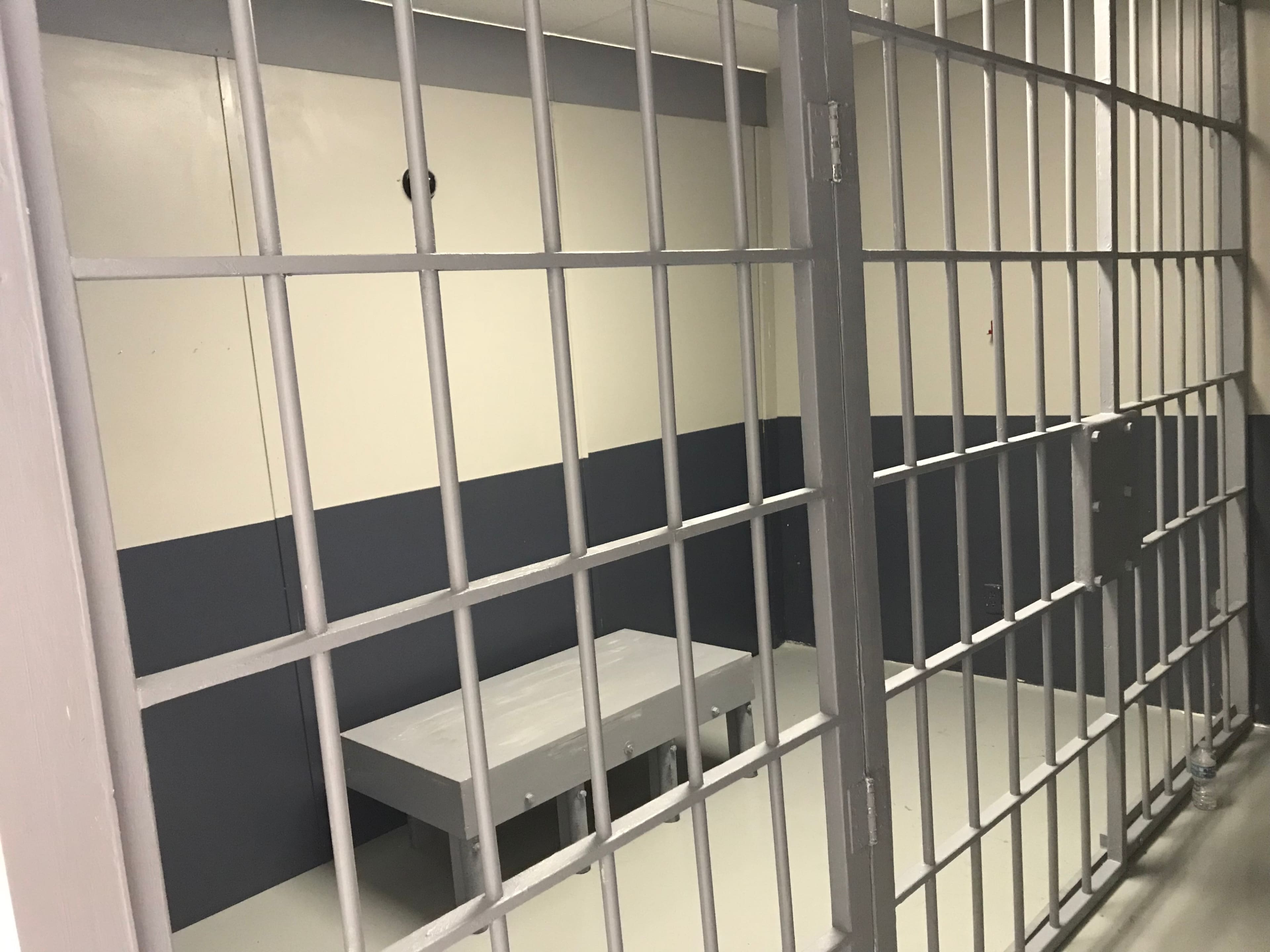 The image shows a jail cell with metal bars and a gray wall, featuring a small table in the center of the room.