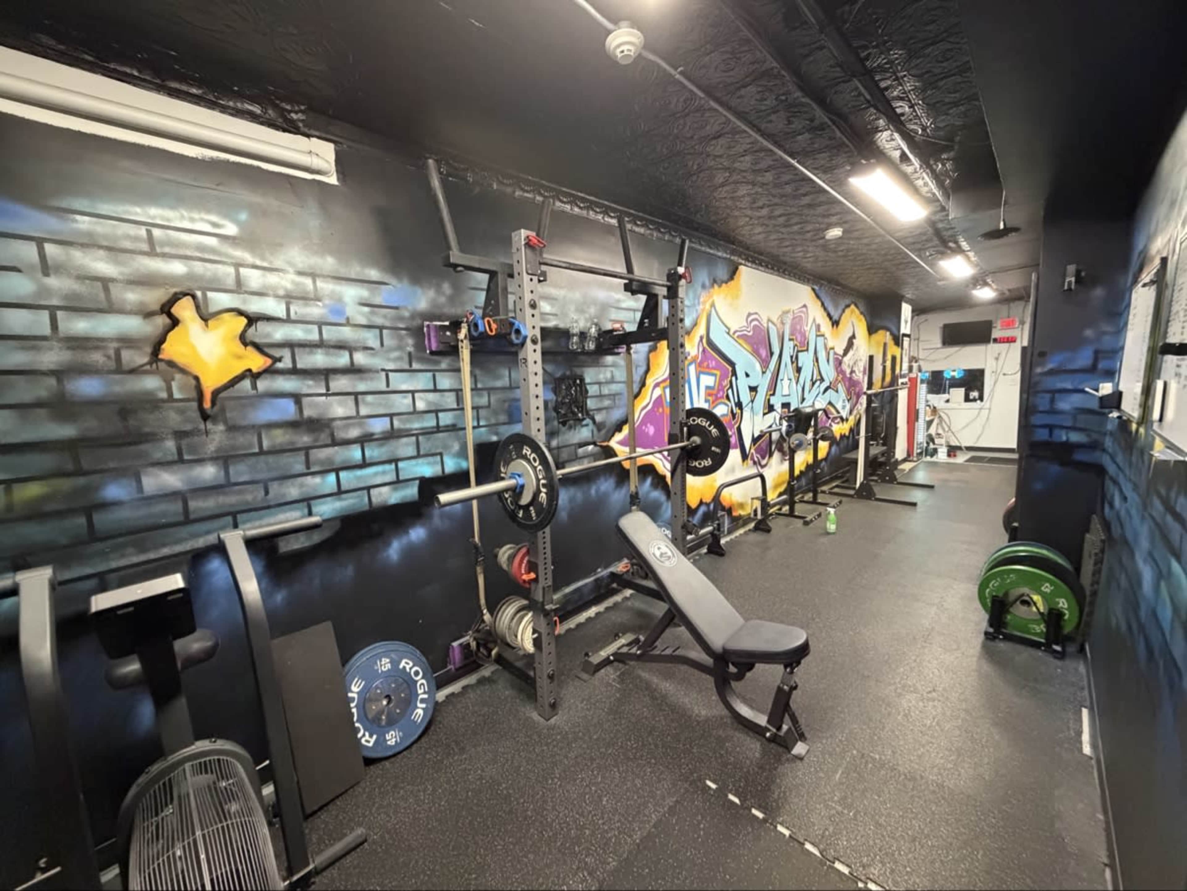 The image shows a gym interior featuring weightlifting equipment, a bench, and a colorful graffiti mural on the wall.