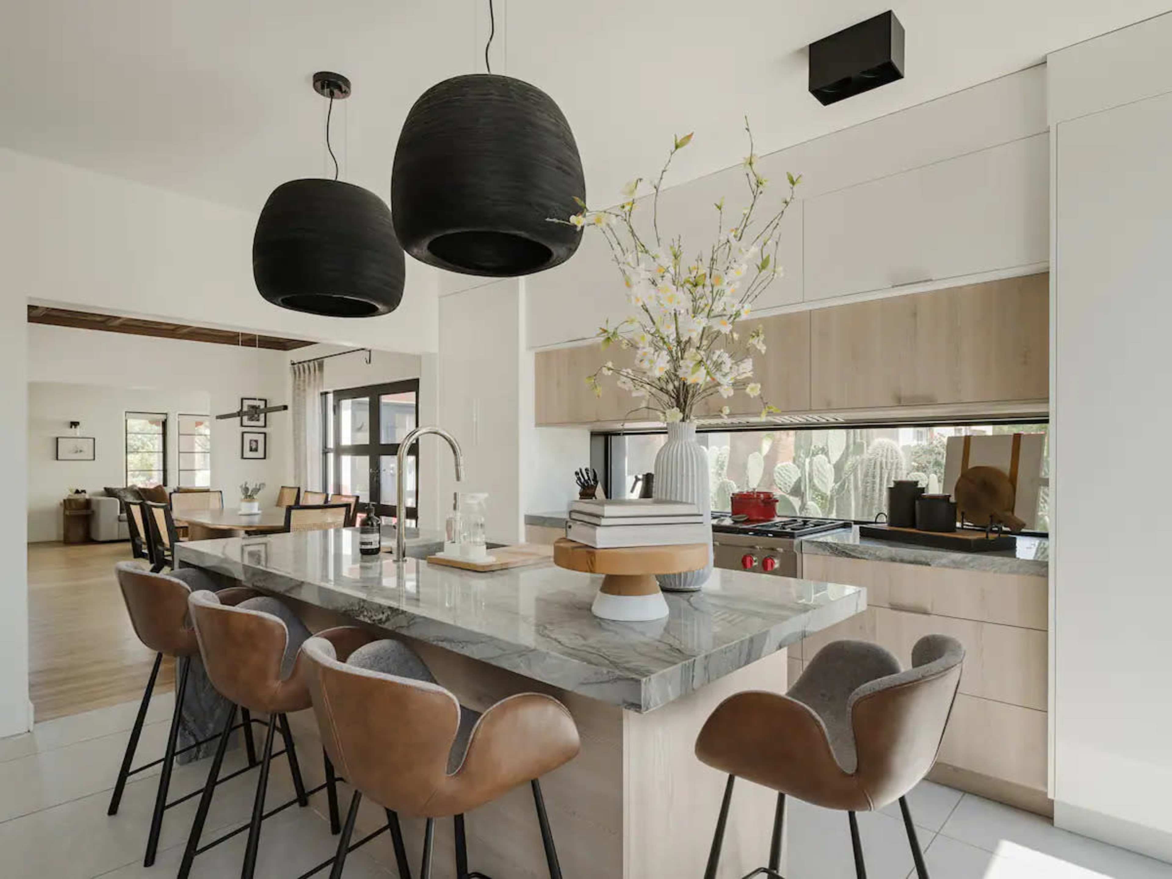 A modern kitchen features a large island with a marble countertop, four brown upholstered chairs, and pendant lights above.