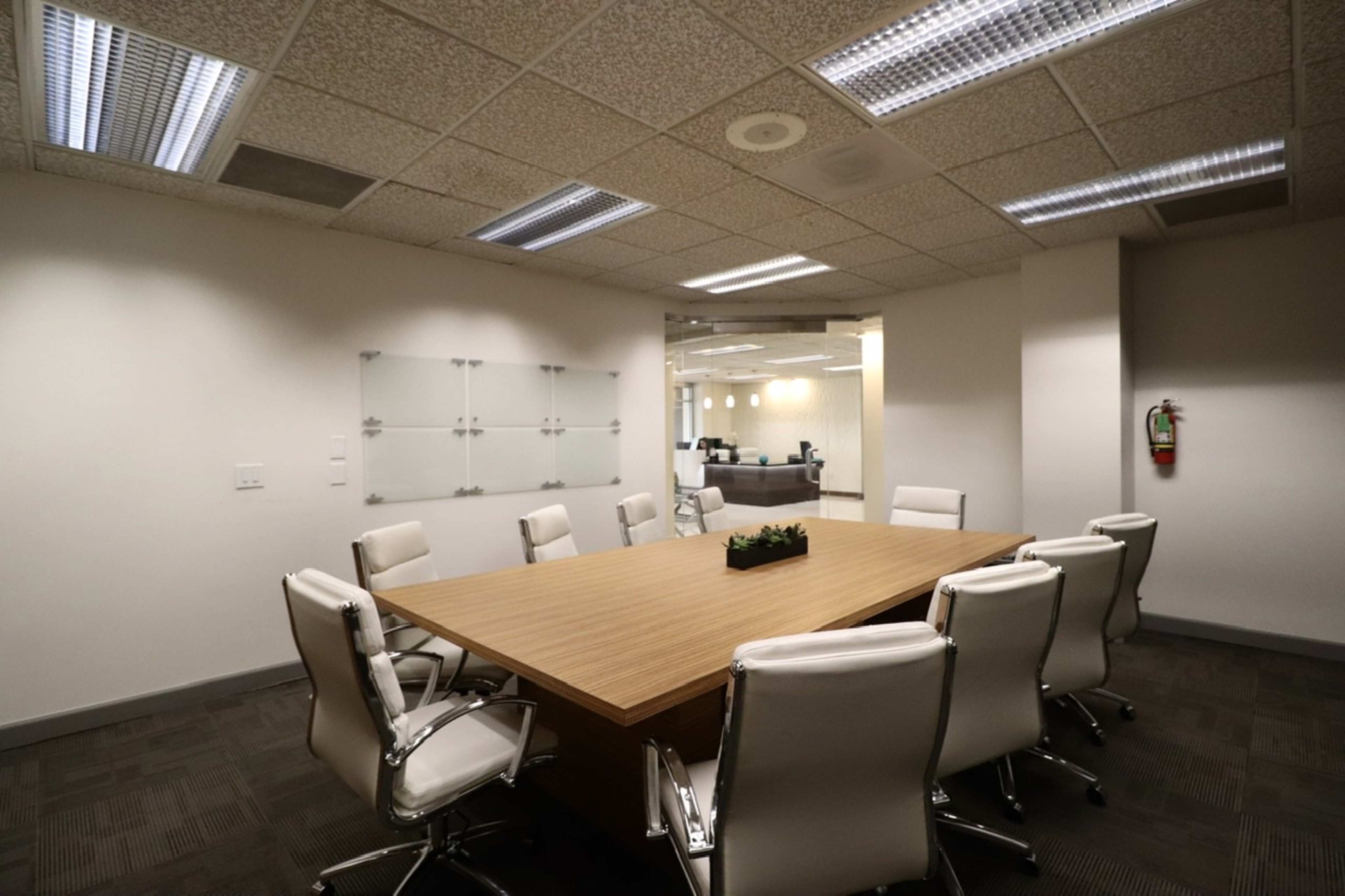 A conference room features a large wooden table surrounded by eight white chairs, with glass walls and a fire extinguisher visible in the background.