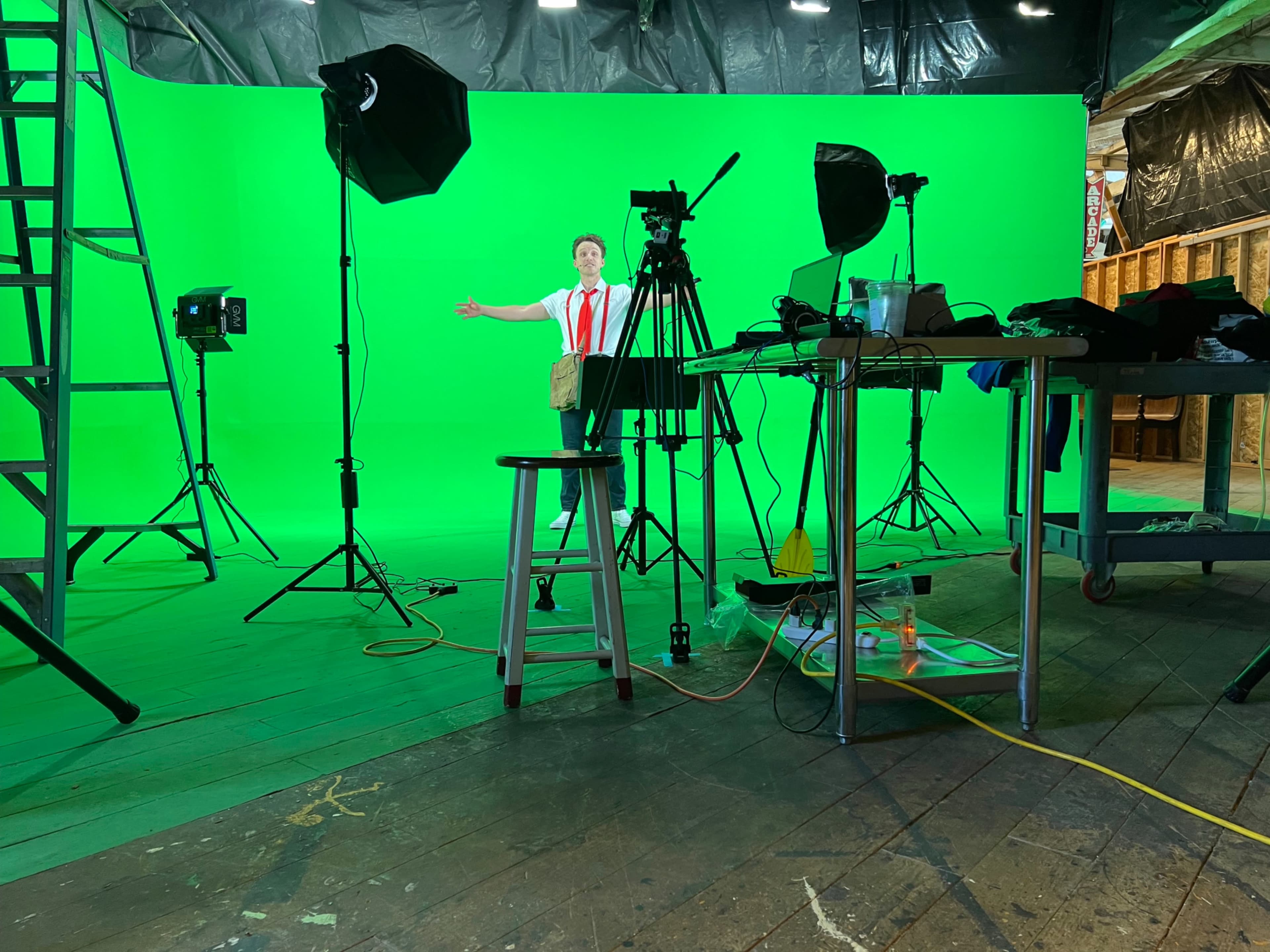A person stands in front of a green screen with filming equipment and lighting set up around them in a studio.