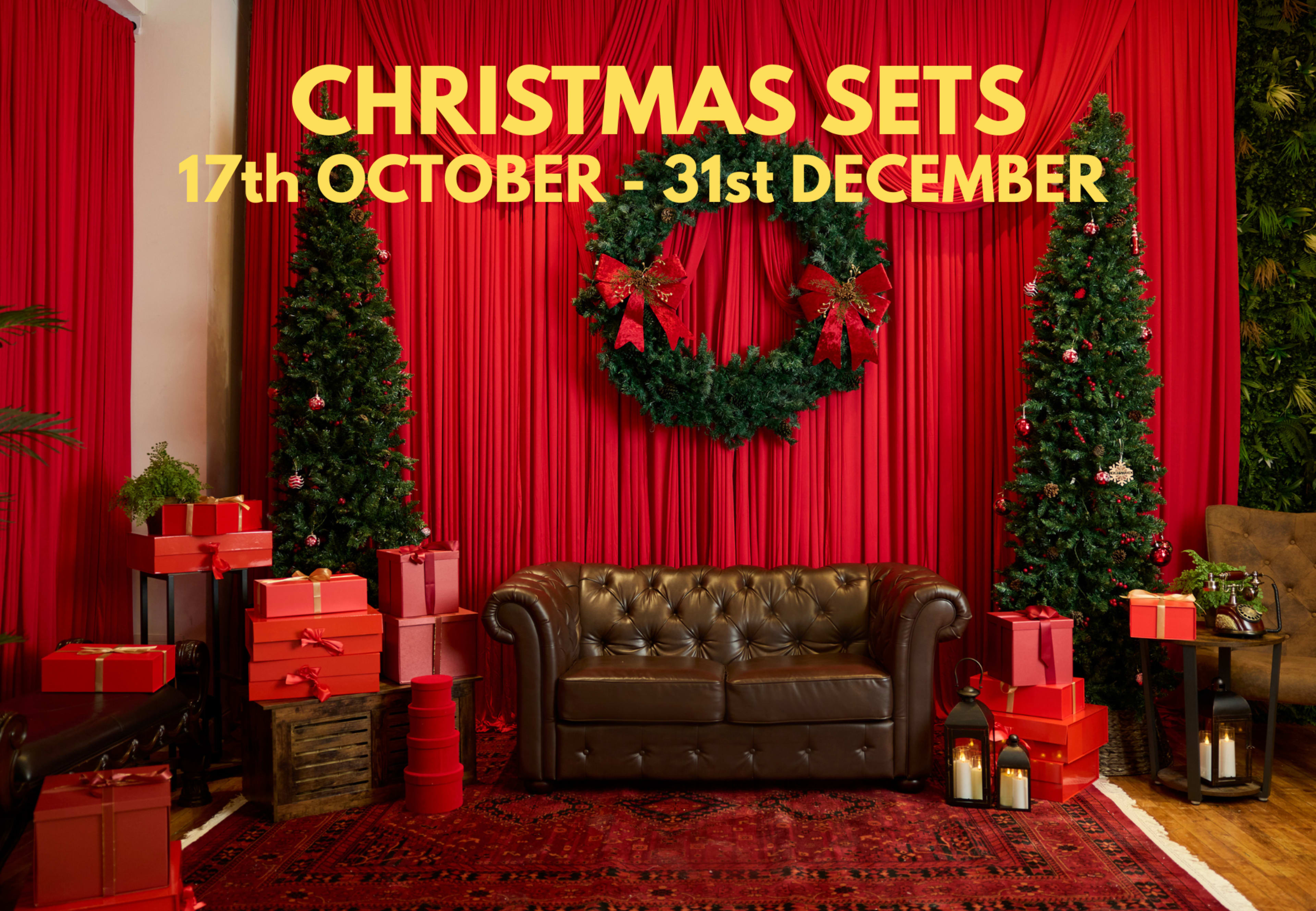 The scene features a festive Christmas-themed setting with a red backdrop, a green wreath, two decorated trees, a brown leather sofa, and numerous wrapped gifts.