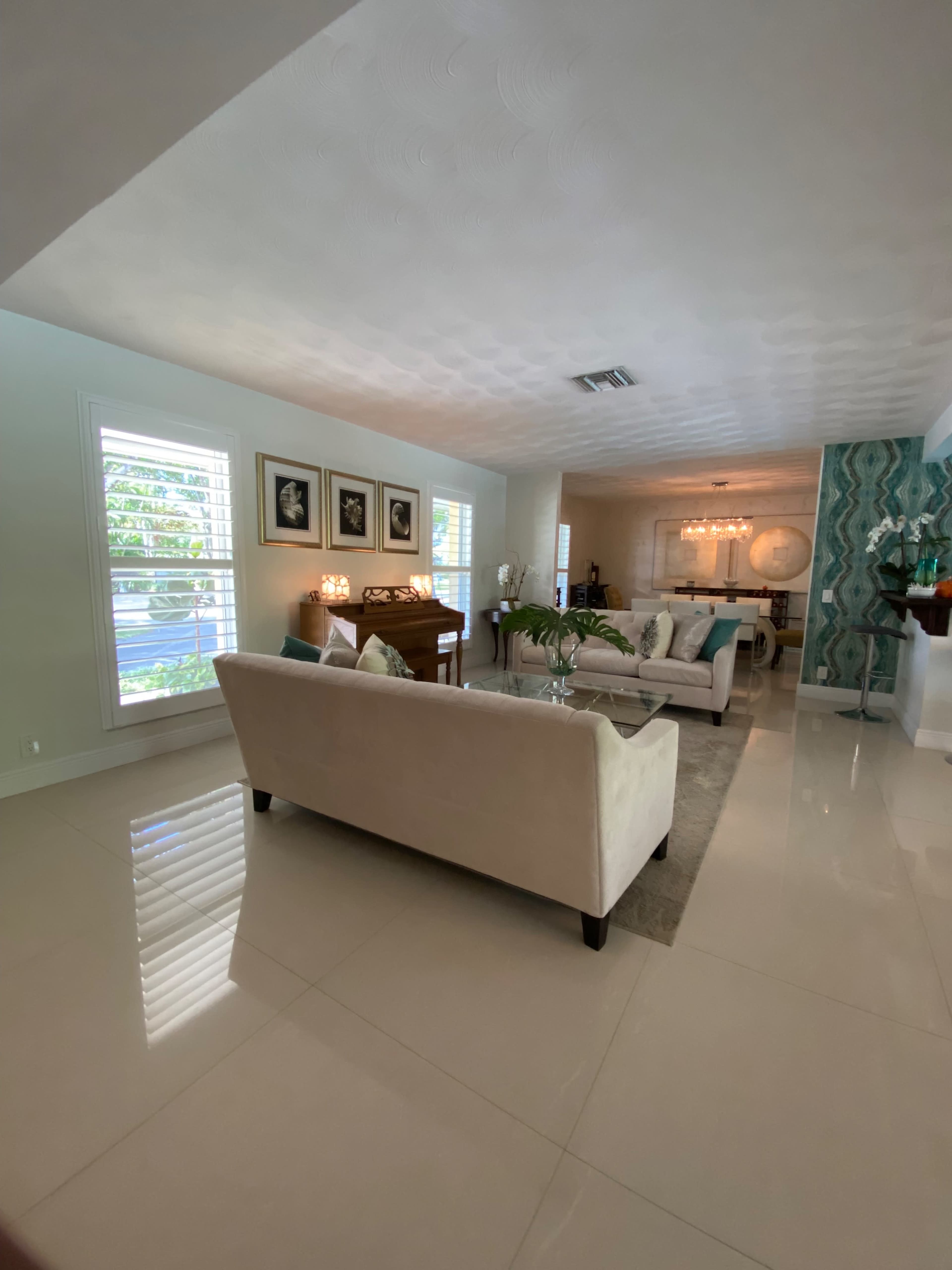The image shows a modern living room with white furniture, polished tile floors, and large windows allowing natural light to enter.