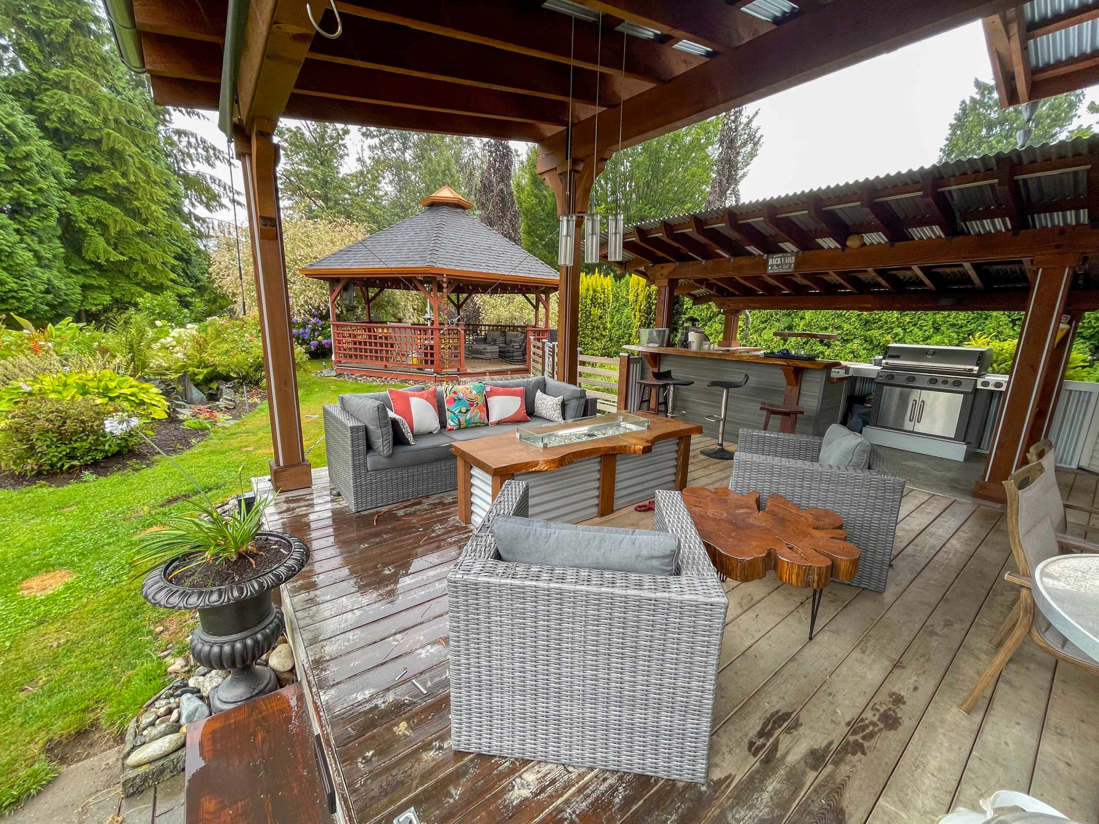 A covered outdoor patio area with seating arrangements, a fire table, and a barbecue station, surrounded by lush greenery.