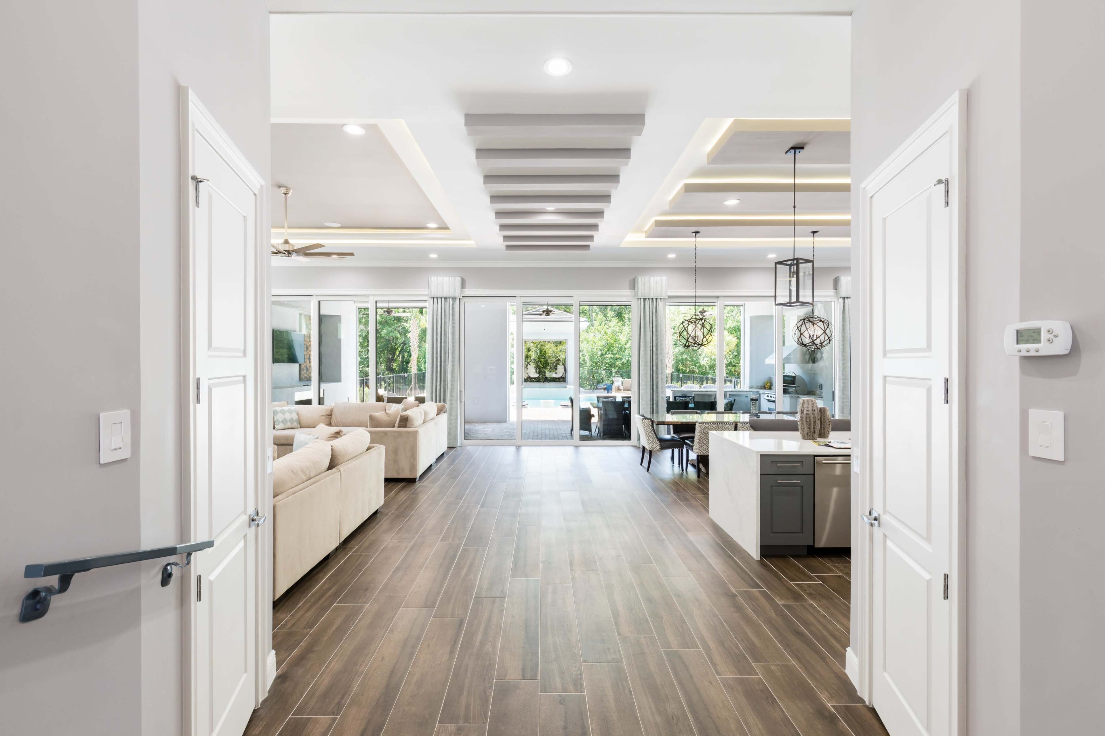 The image shows a modern interior space featuring an open floor plan with a view of a living area and kitchen, illuminated by natural light from large windows.