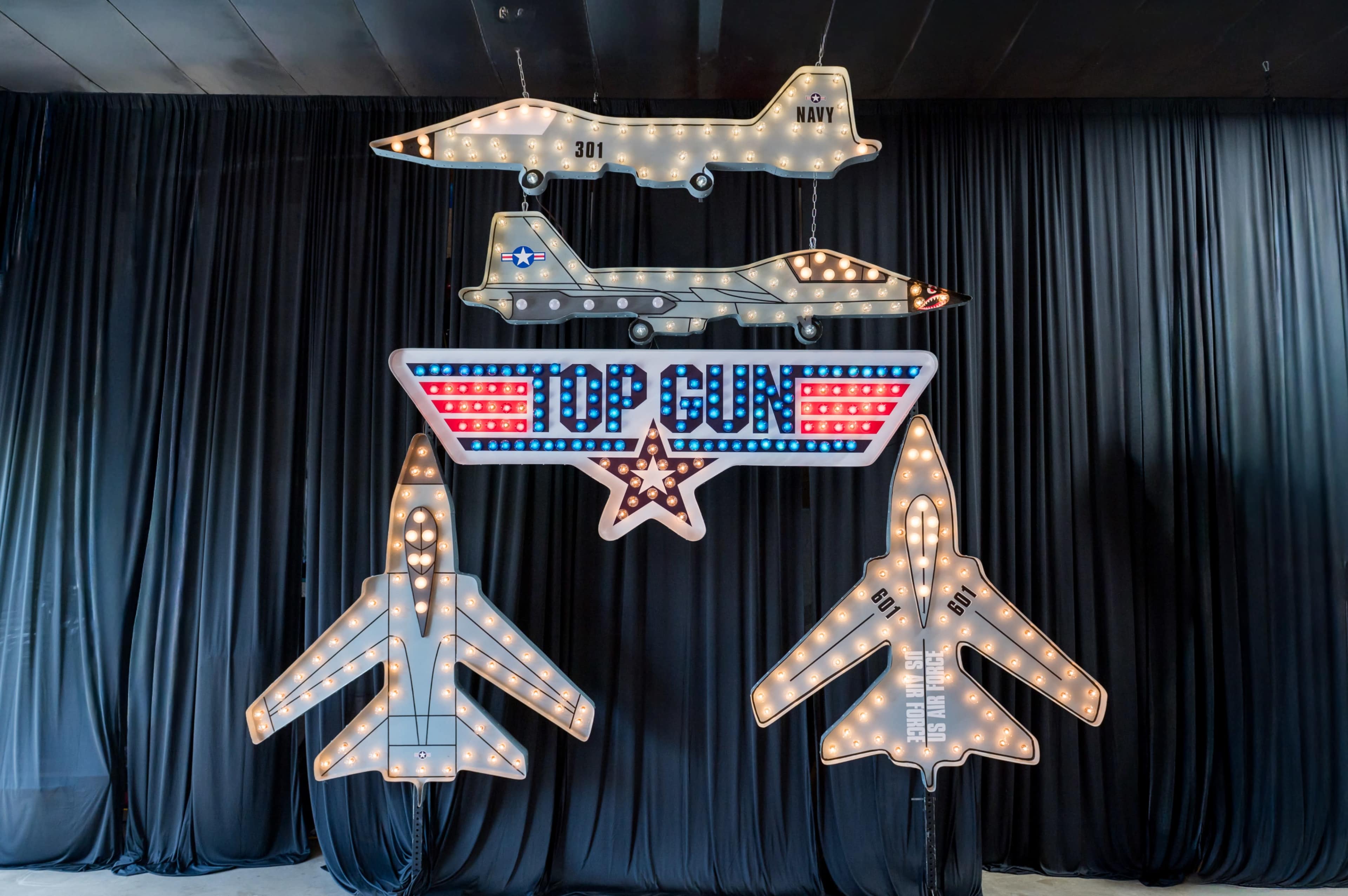 The image features illuminated model aircraft and a "TOP GUN" sign hanging against a black curtain backdrop.
