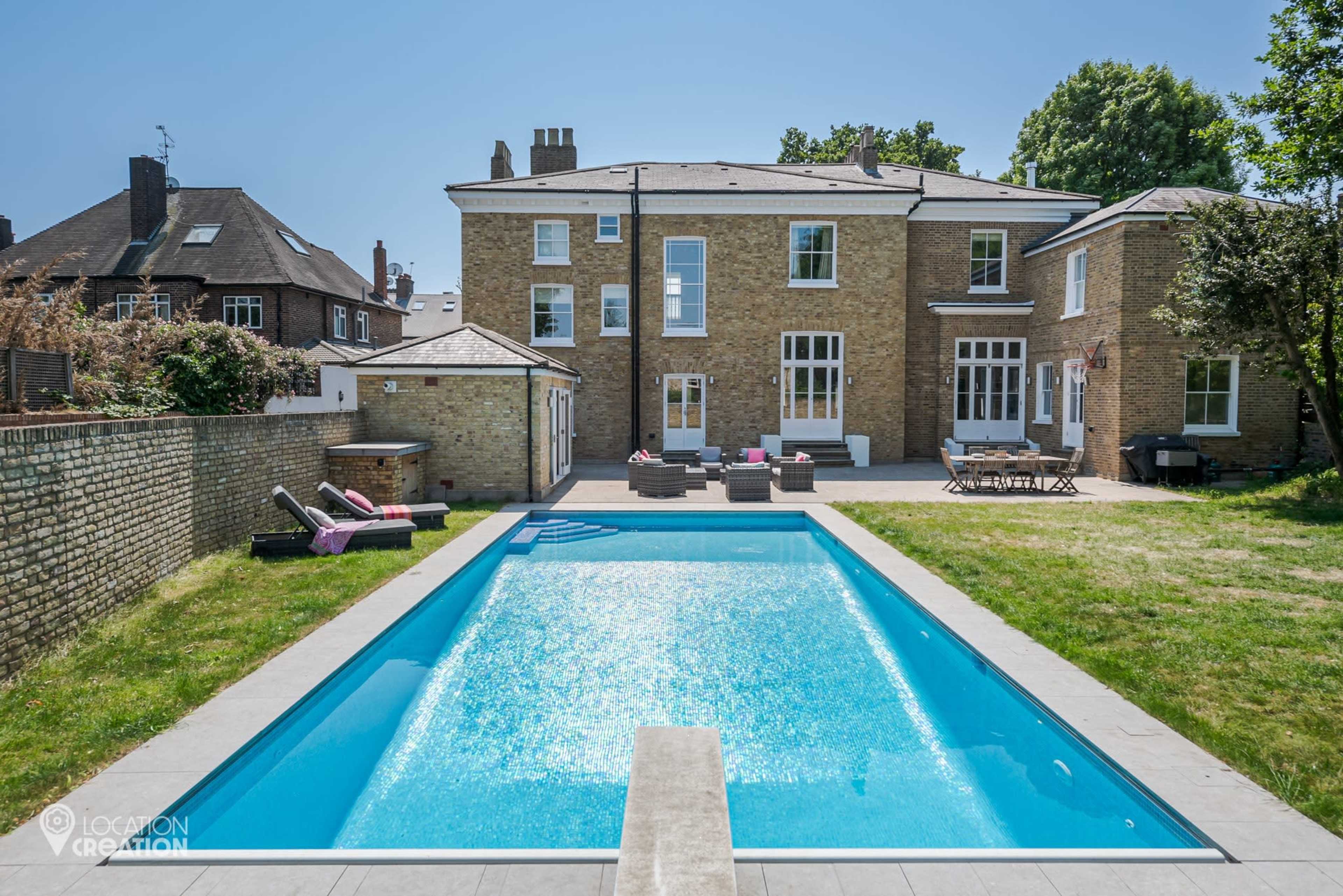 The image shows a modern two-story brick house with a swimming pool in the foreground and a grassy area in the background.