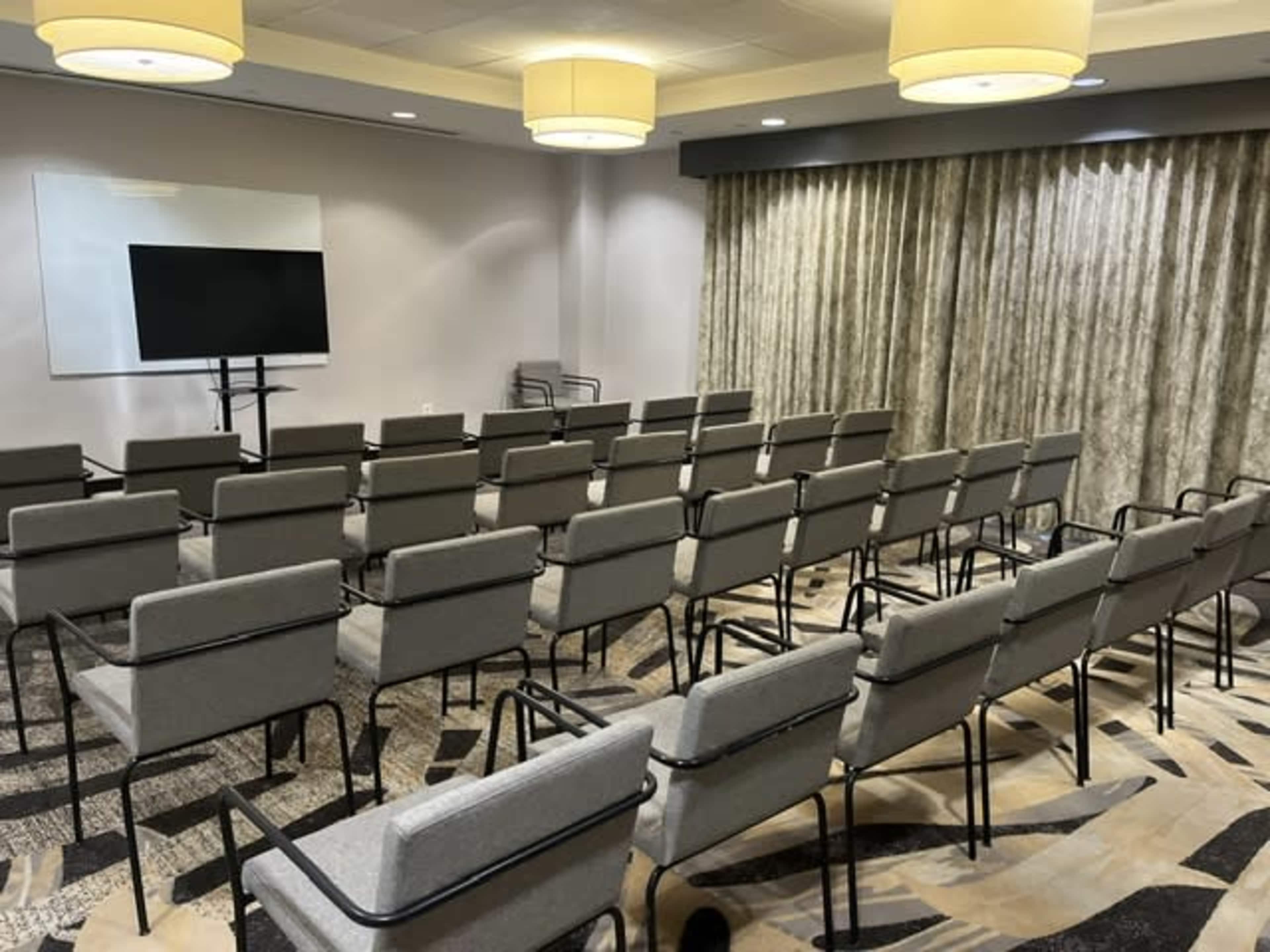 The image shows a conference room arranged with rows of gray chairs facing a wall-mounted screen and illuminated by overhead light fixtures.