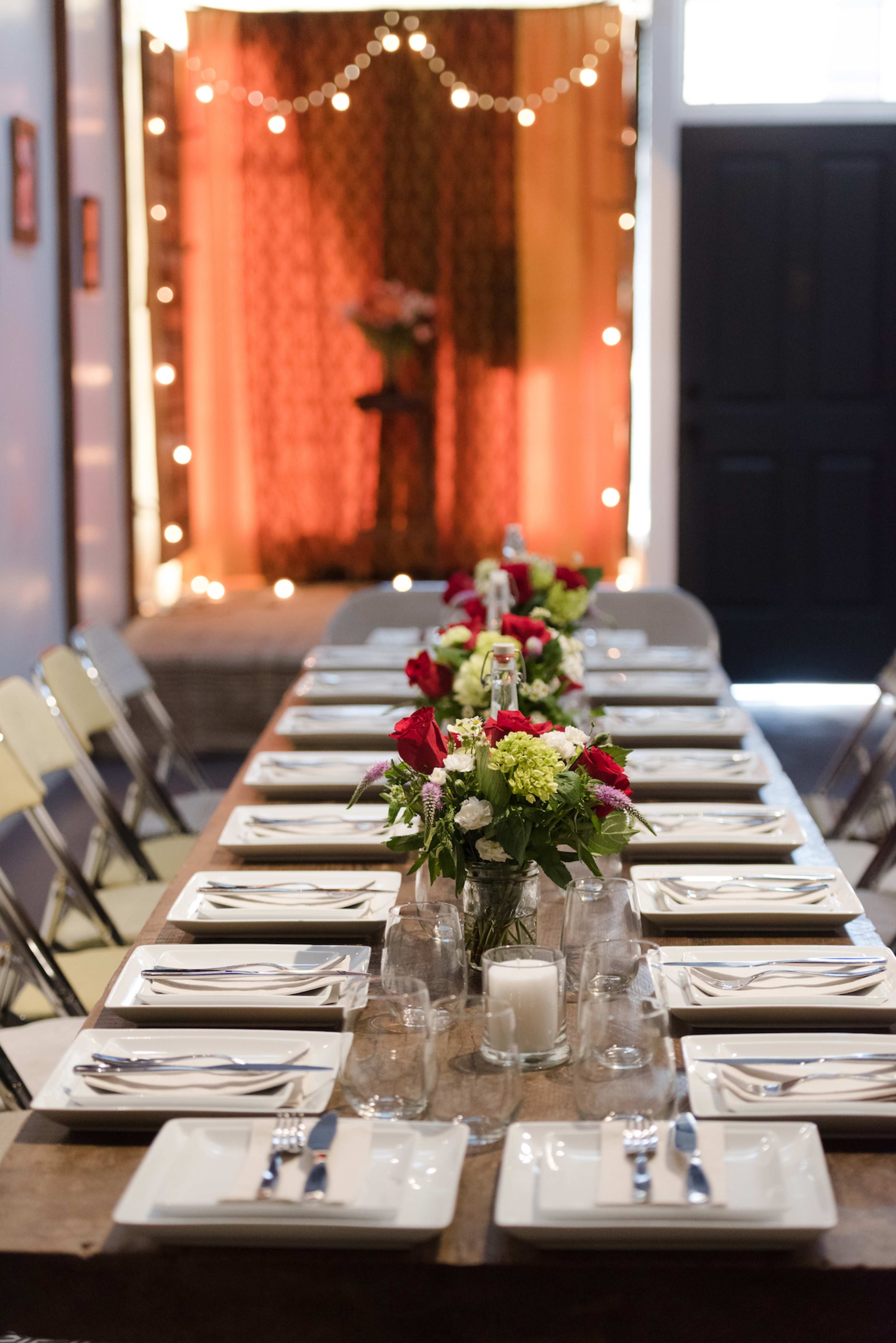 A long table is set with white plates, cutlery, and centerpieces of flowers, with a backdrop of string lights and a decorative curtain in a softly lit room.