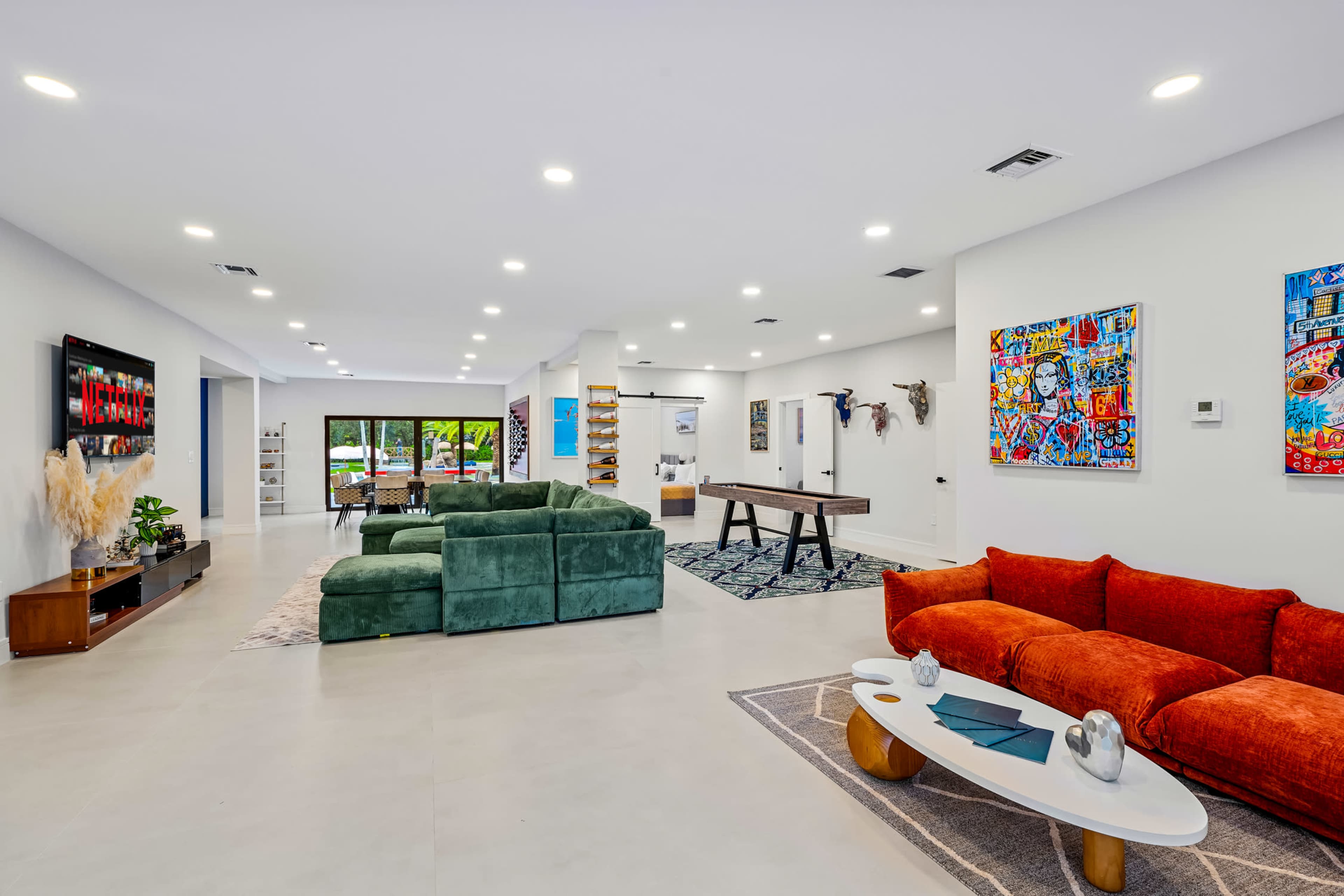 The image shows a spacious, well-lit living area with a green sectional sofa, a red couch, a dining table, and decorative wall art.