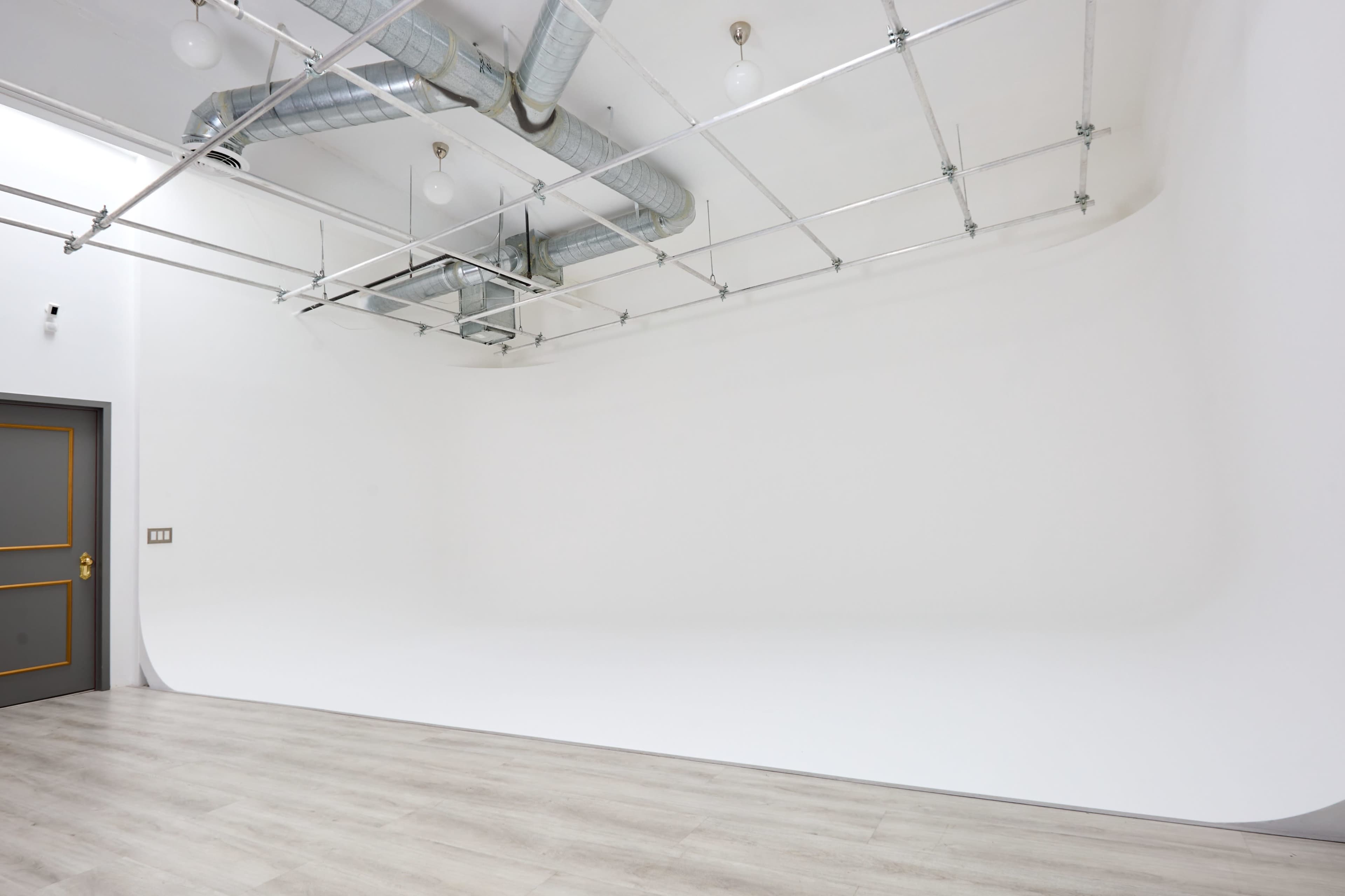 The image shows a minimalist white studio space with a seamless backdrop and exposed ductwork on the ceiling.
