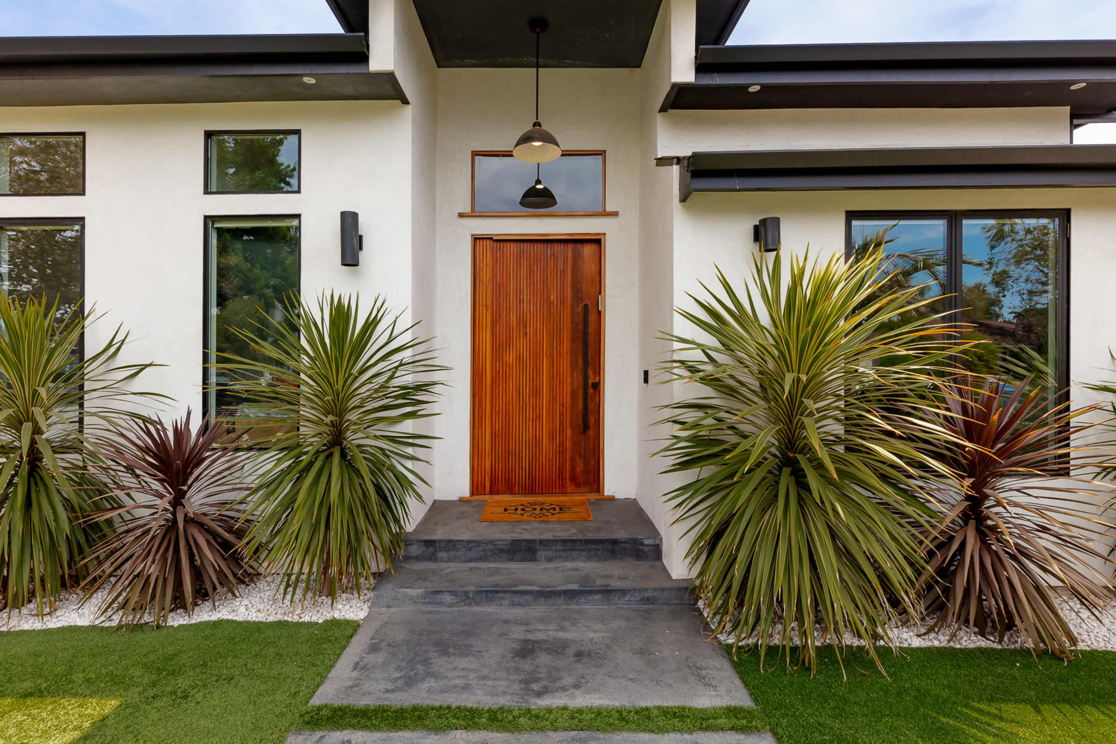 The image shows a modern house entrance with a wooden door, flanked by tall, spiky plants and a neatly trimmed lawn.