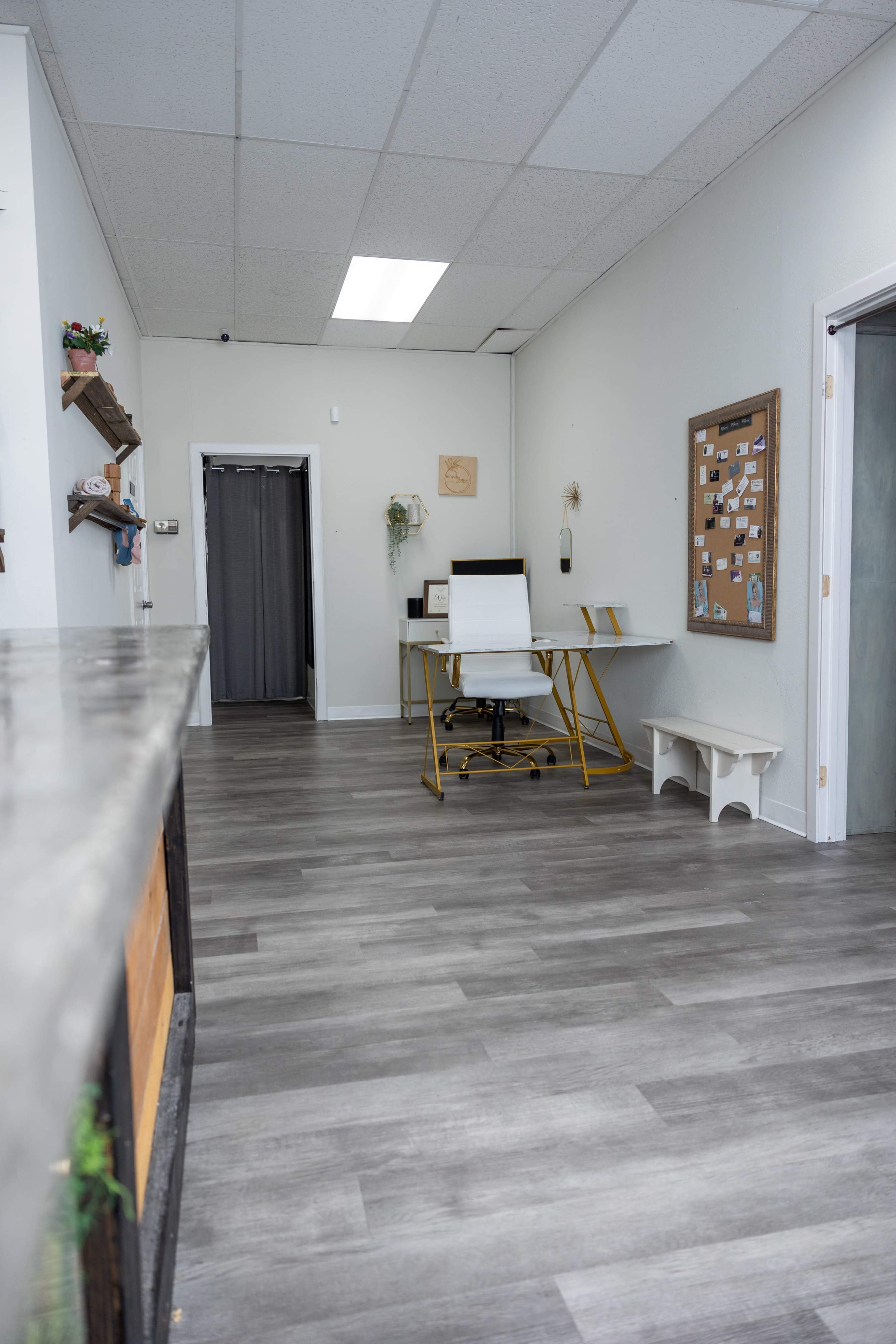 A narrow office interior with a desk and chair at one end, a bulletin board on the wall, and a grey wood floor that extends throughout the space.