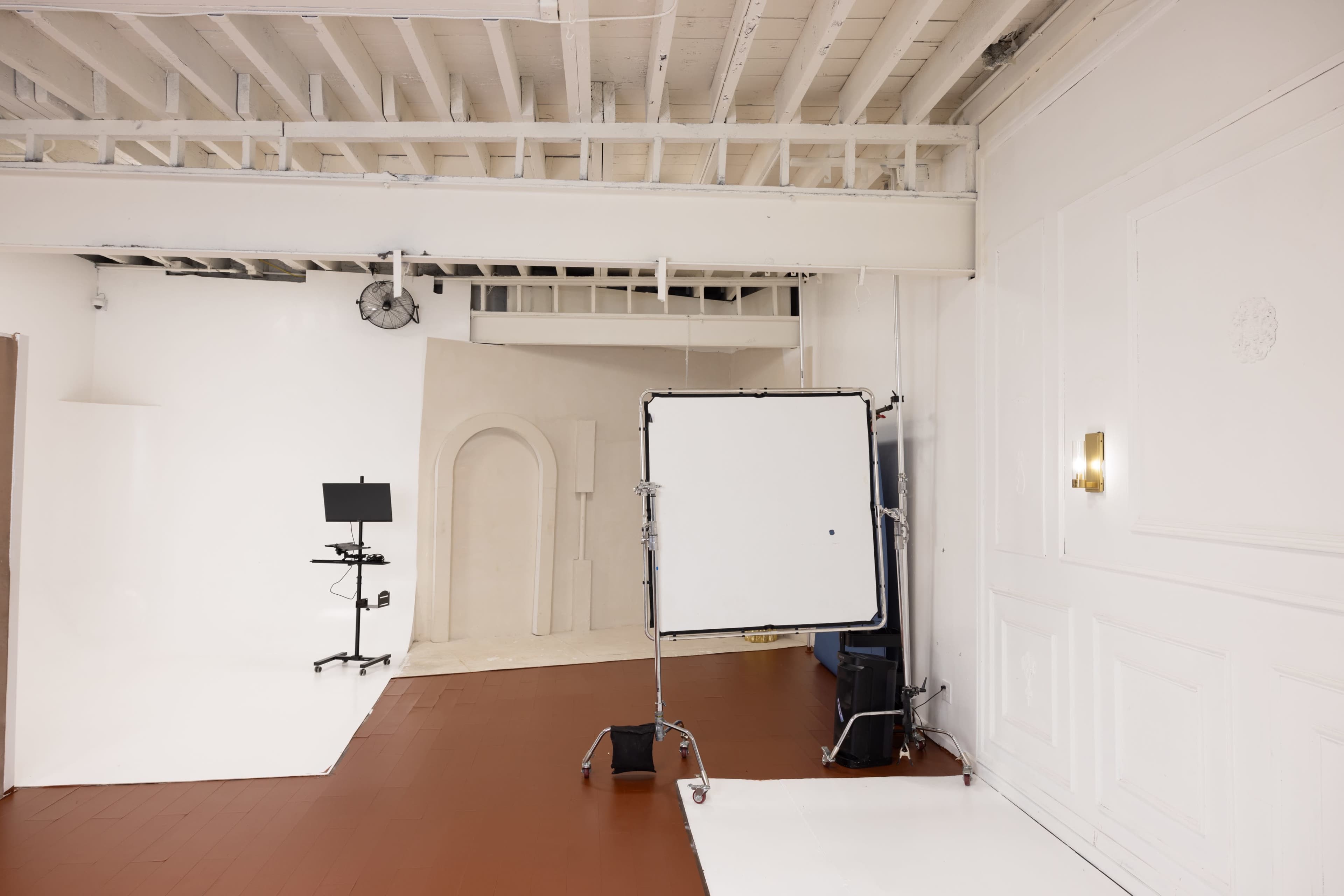 A minimalist photography studio with a white backdrop, a large reflector on a stand, and a monitor on a wheeled cart.