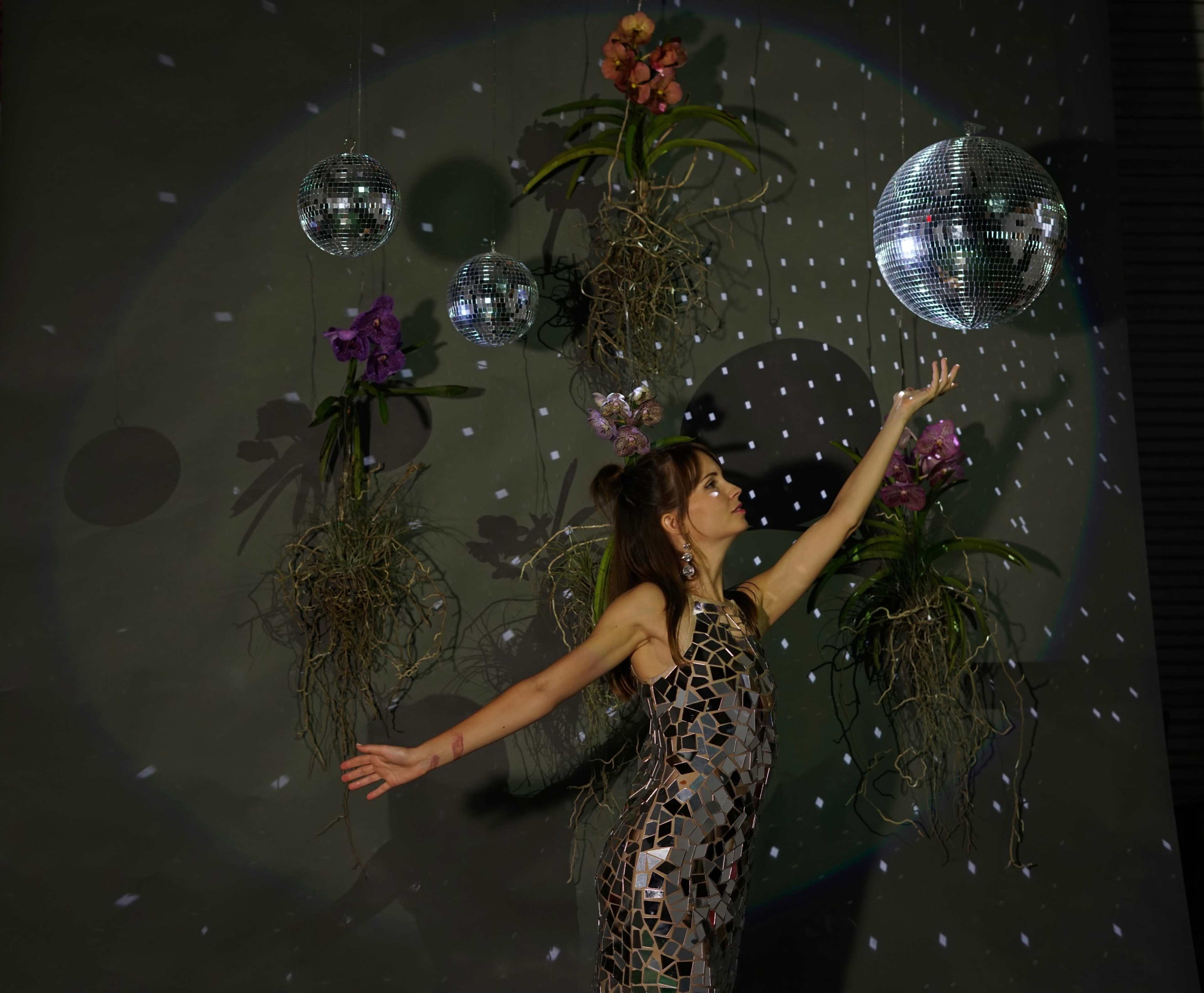 A woman in a mirrored outfit reaches toward a hanging disco ball surrounded by plants and flowers.