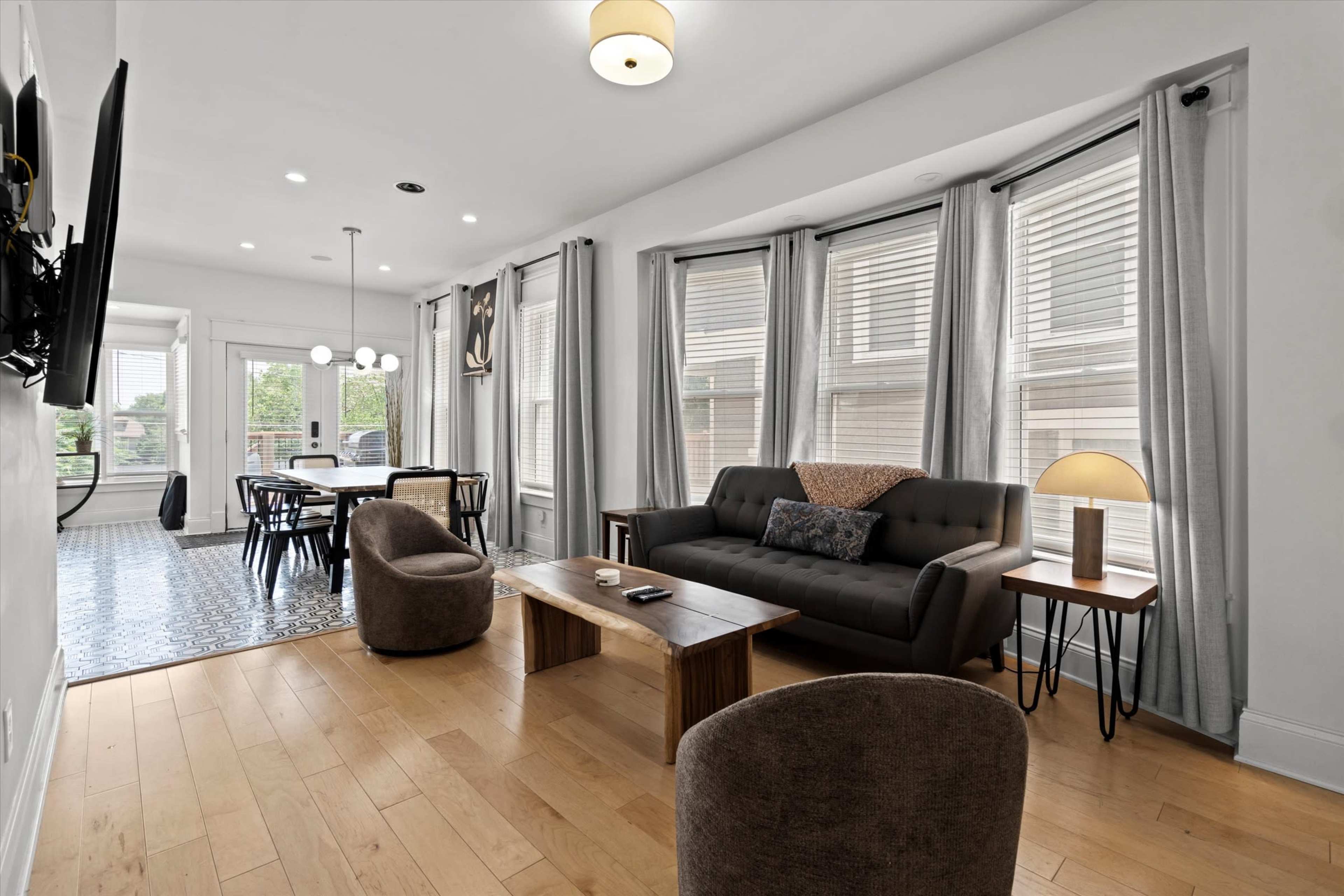 A modern living room features a dark sofa, a wooden coffee table, and two brown chairs, with large windows allowing natural light to brighten the space.