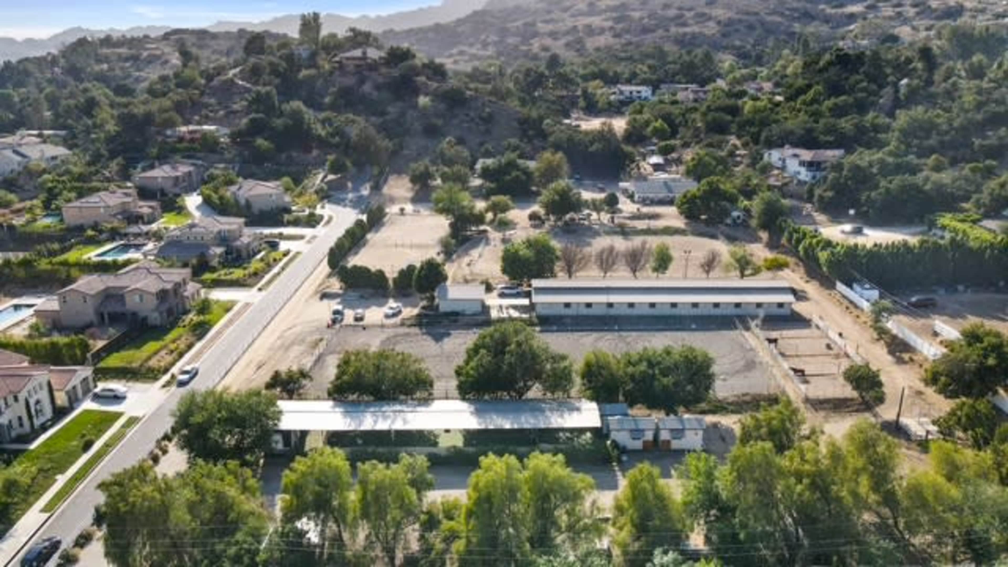 The image shows an aerial view of a residential area with several houses, a wide street, and a section of land with construction or development in progress, surrounded by hills and greenery.