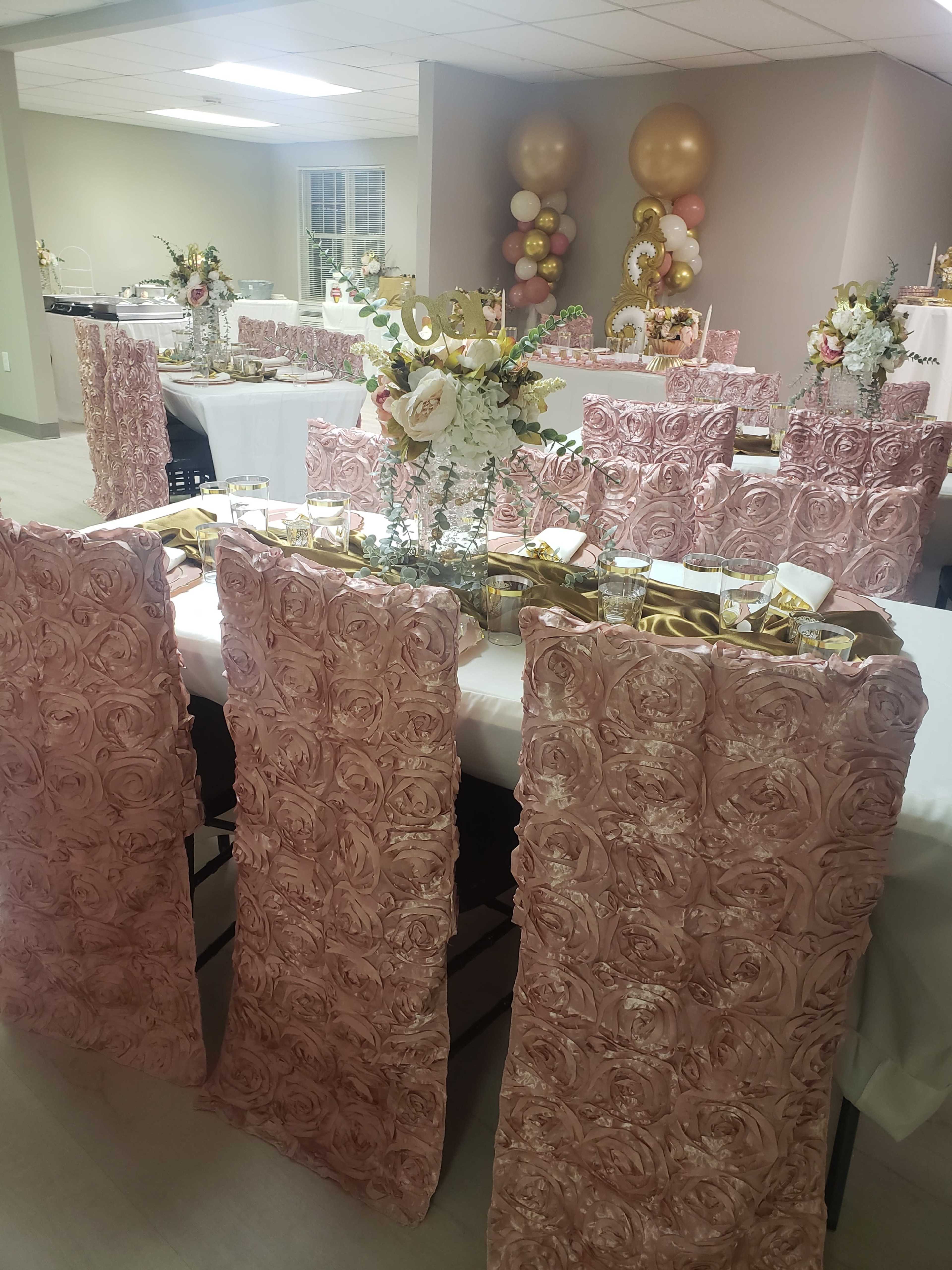 The image shows a banquet room set up for an event, featuring tables with elaborate floral decorations and rose-patterned chair covers in a soft pink hue.