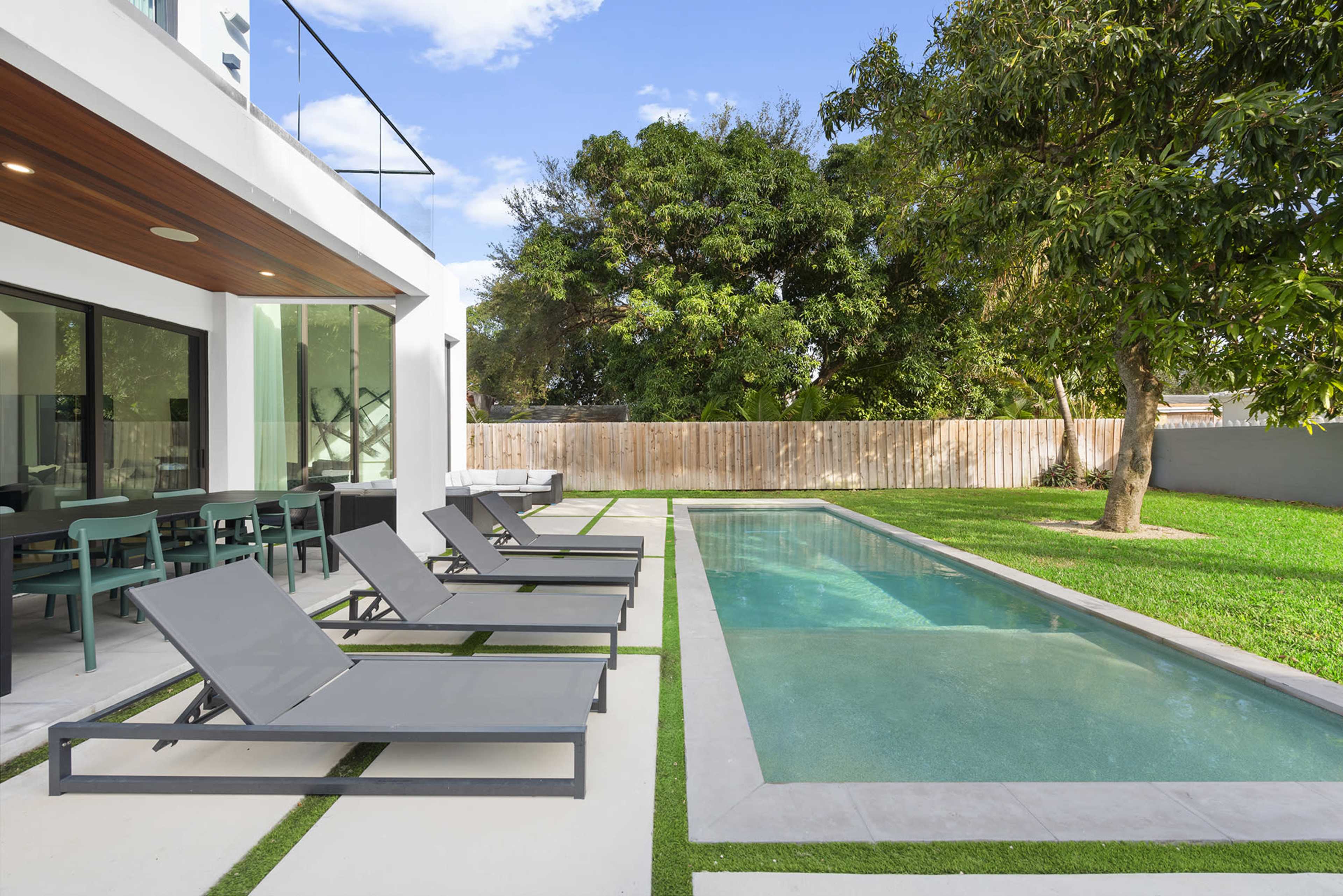 A modern outdoor space features a rectangular swimming pool alongside gray lounge chairs and a dining area, framed by green grass and trees.