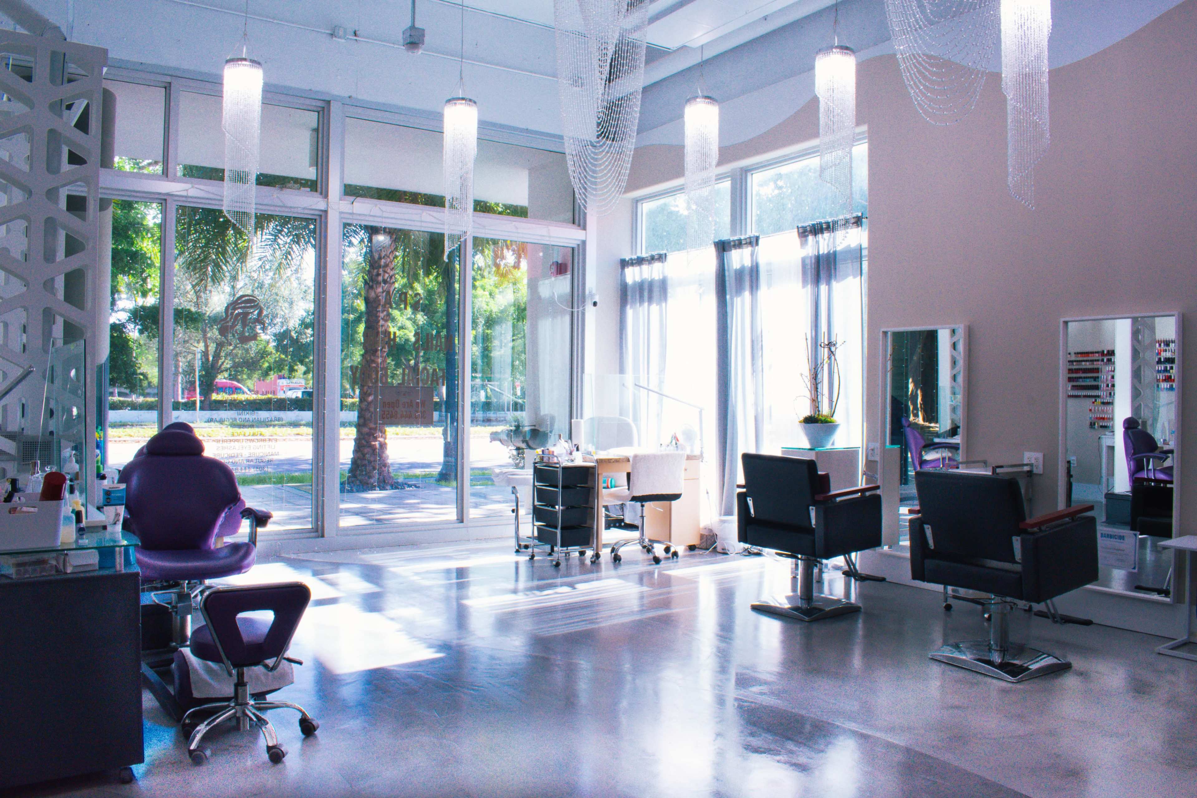 A modern salon interior with two styling chairs, a manicure station, and large windows allowing natural light to enter.