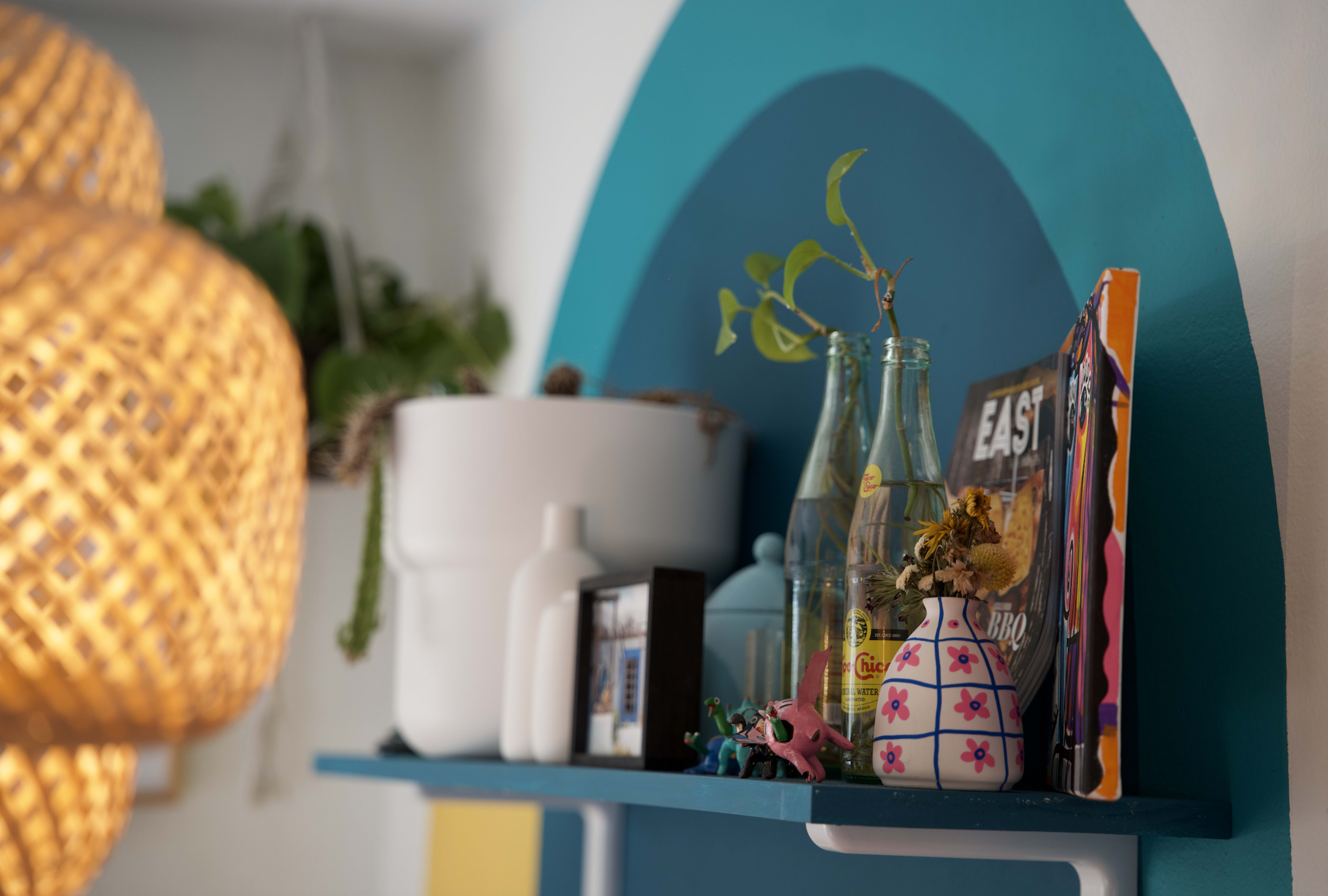 A shelf displays a collection of decorative items, including plants, glass bottles, and magazines, against a blue and white backdrop.