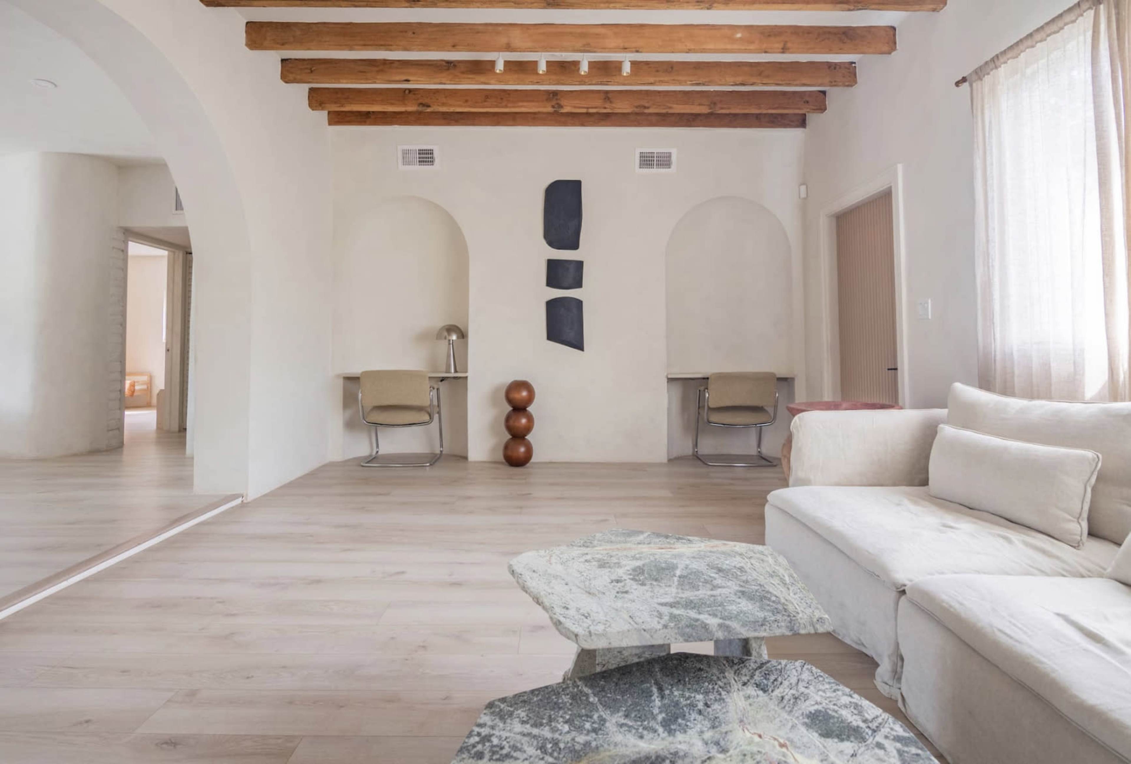 A spacious room with wooden beamed ceilings, featuring a modern sofa, two abstract wall decorations, and two work desks.