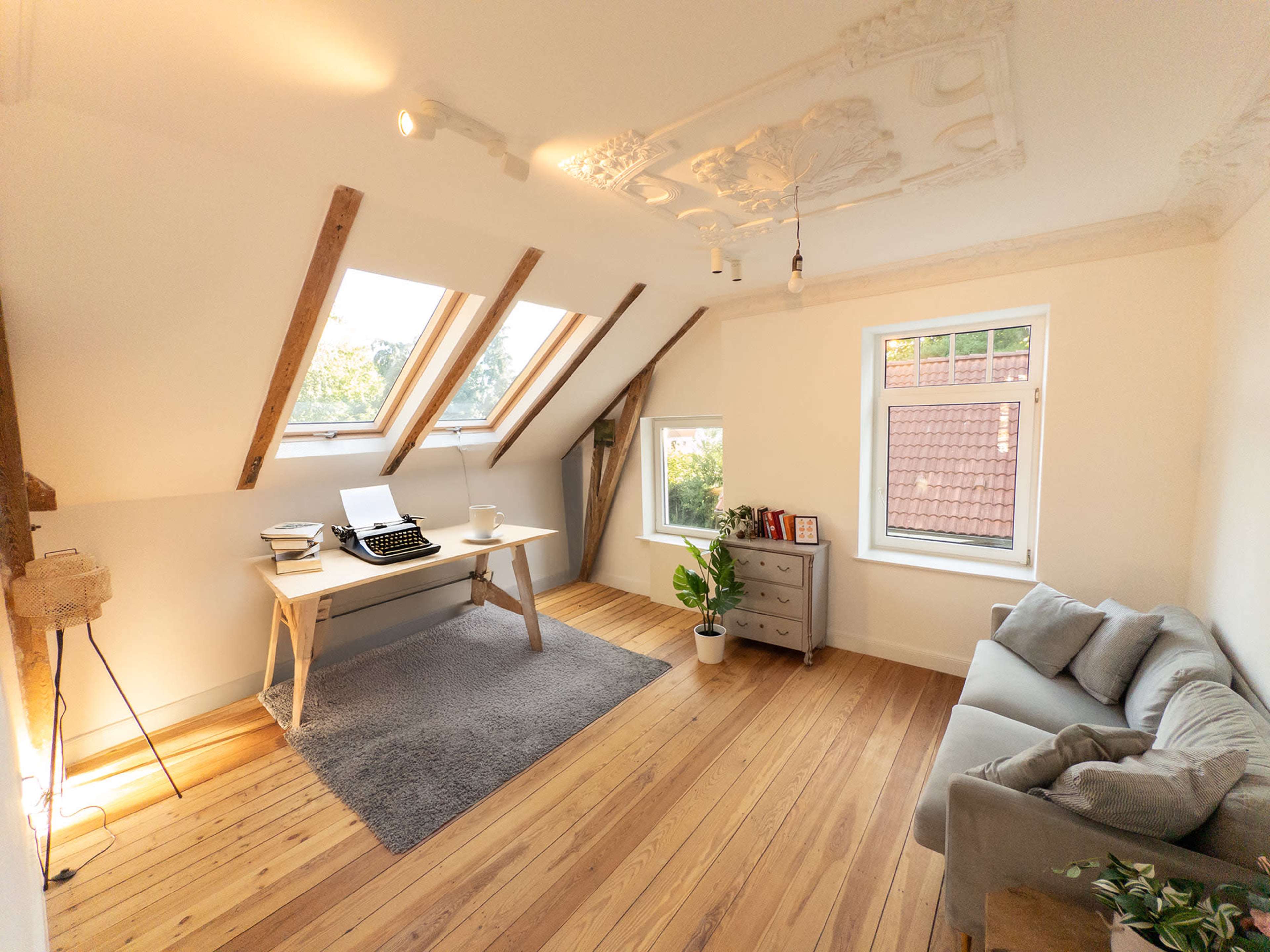 The room features a desk with a typewriter, a gray couch, and a large window, all set against wooden flooring and slanted ceilings.