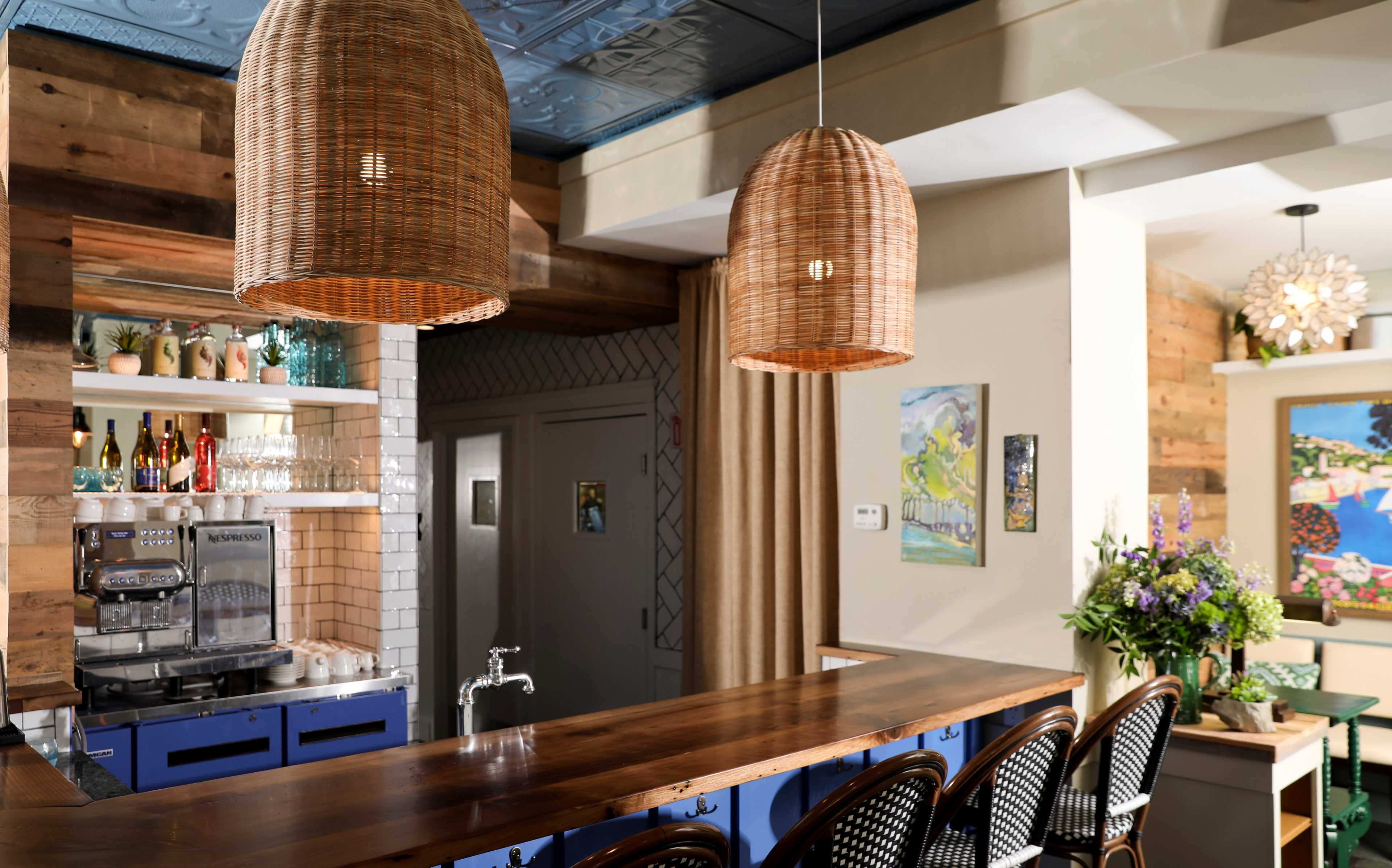 A bar area with a wooden countertop, woven pendant lights, and a shelf displaying various bottles and glassware.