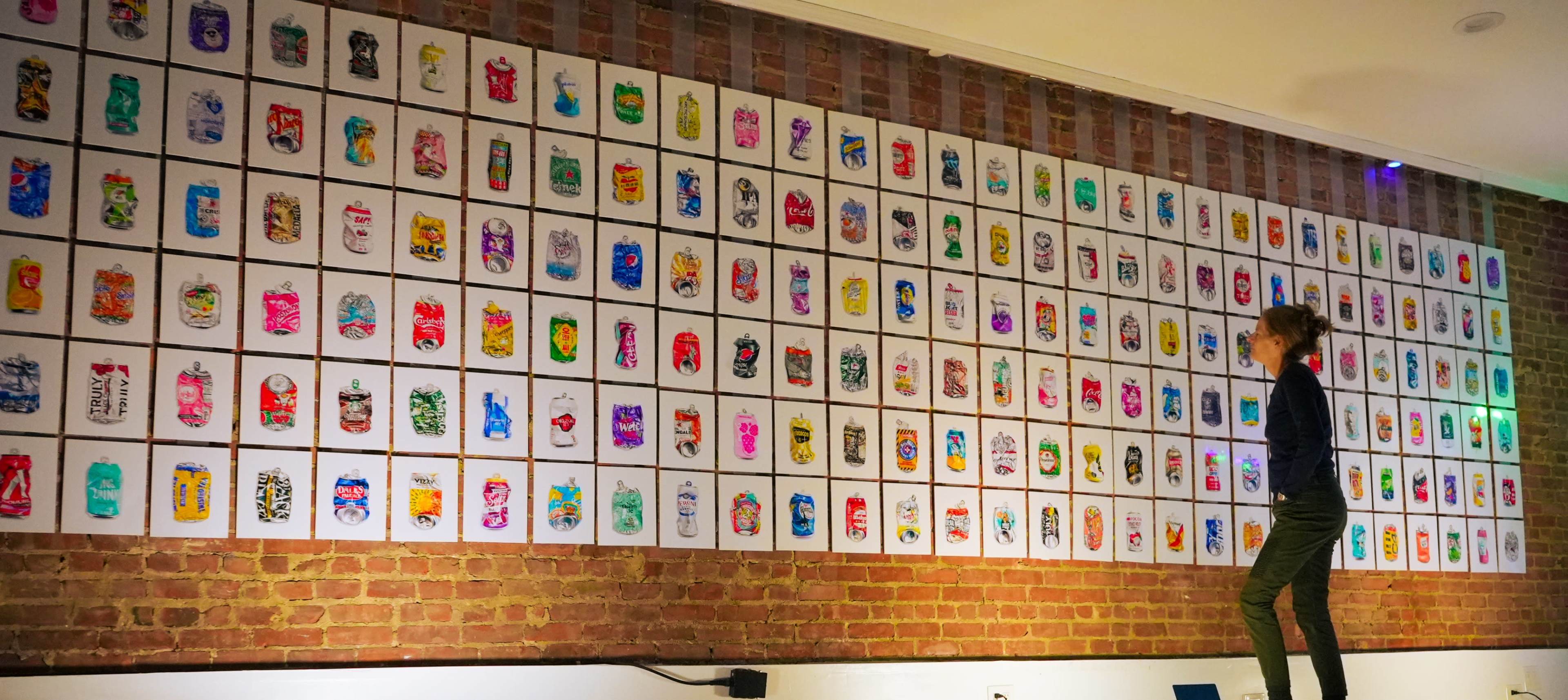 A person stands in front of a wall displaying an extensive collection of colorful artworks arranged in a grid pattern.