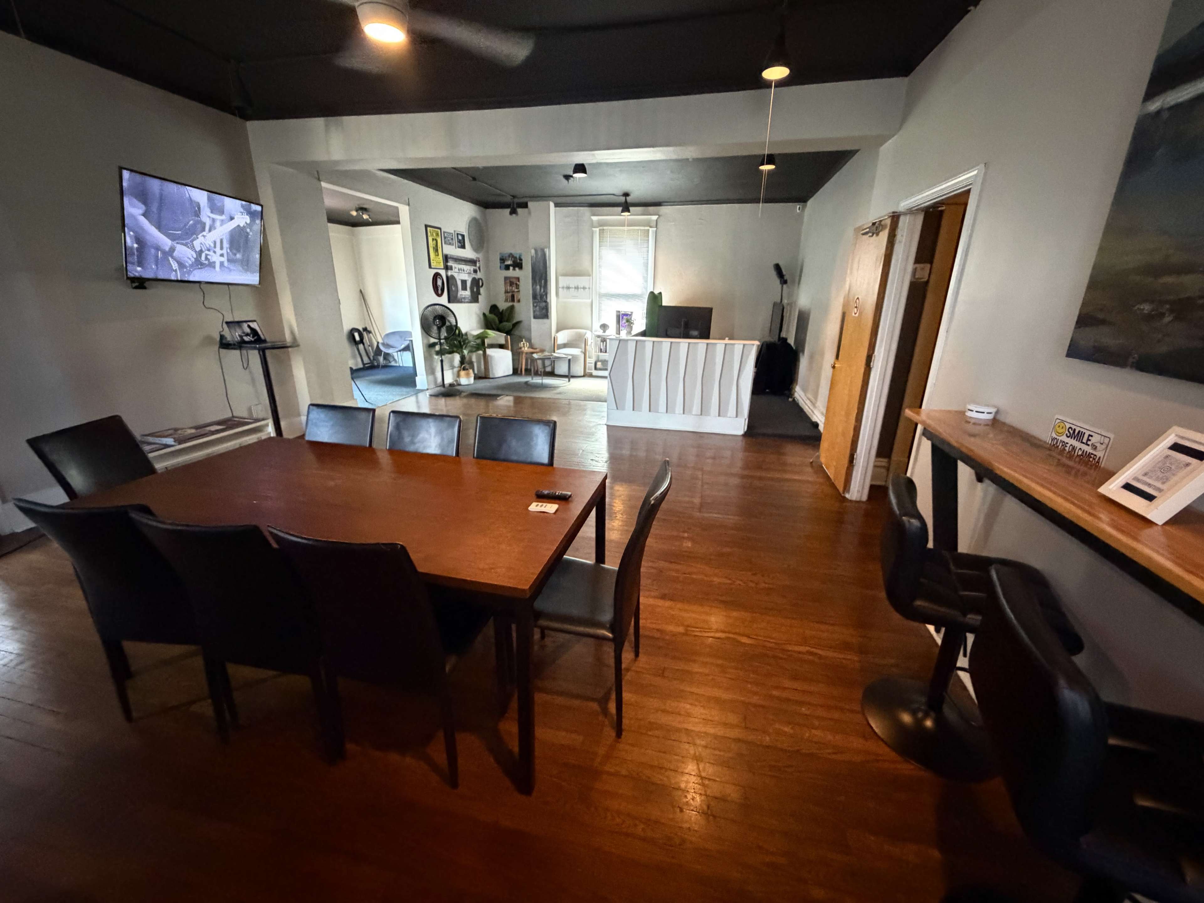 The image shows a spacious room with a large wooden table surrounded by chairs, a TV mounted on the wall, and a separate area with seating and a reception desk.